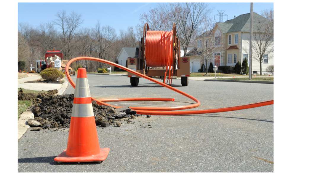 Street repair work with orange traffic cone, construction hoses, and a large truck in a neighborhood with houses and leafless trees.