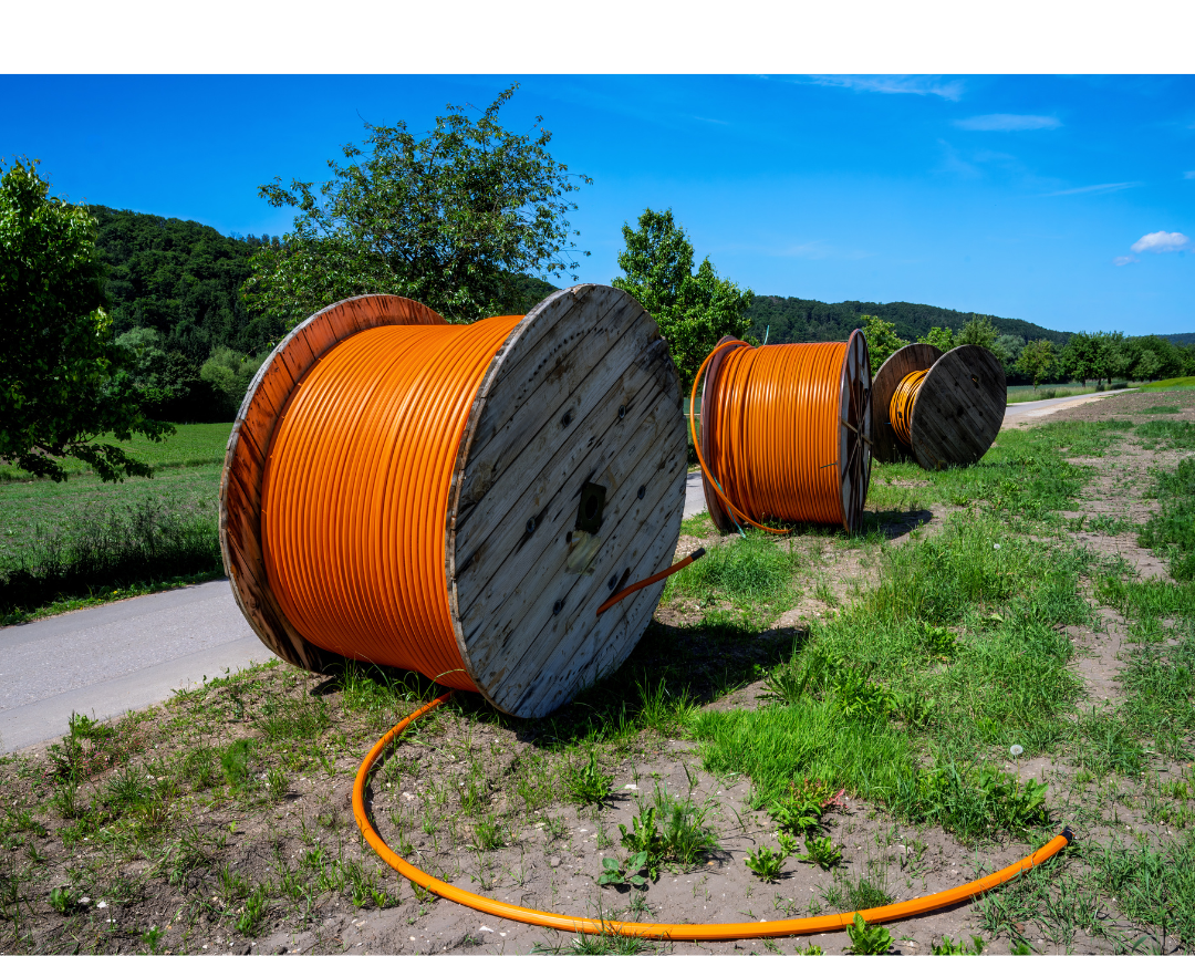 Three large wooden spools with orange cable on grassy roadside against green trees and hills, under blue sky.