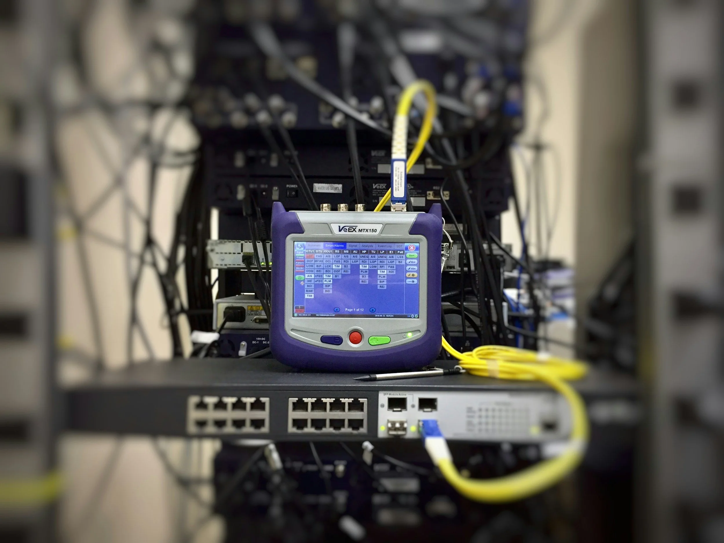 Close-up of a network rack with various cables and a network testing device showing a colorful screen with status indicators.