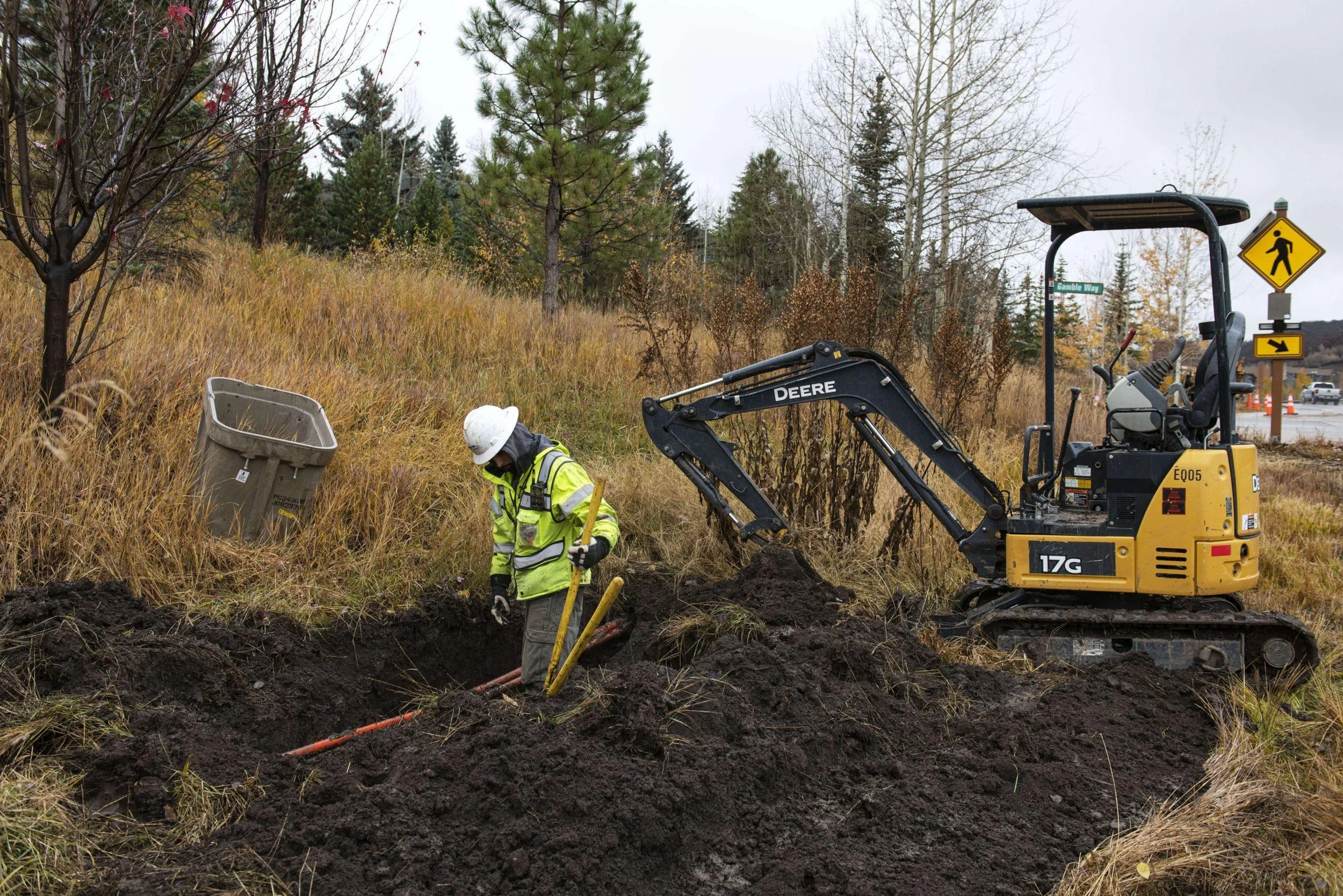 Construction workers digging a trench with a small excavator on the side of a road. One worker uses a shovel while the other stands nearby. There are trees and a street sign in the background.
