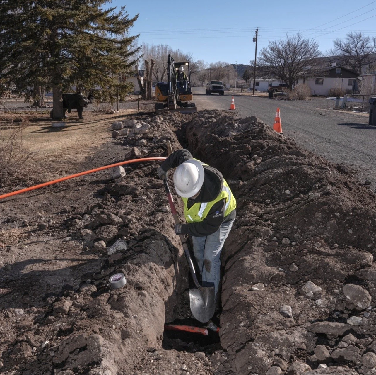 
Worker digging a trench with a shovel in a residential area, with workers and construction equipment in the background.