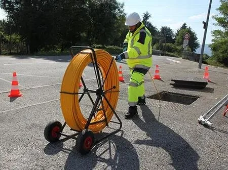 Utility worker in high-visibility clothing and hard hat handling a large coil of orange cable with a reel cart at an outdoor work site.