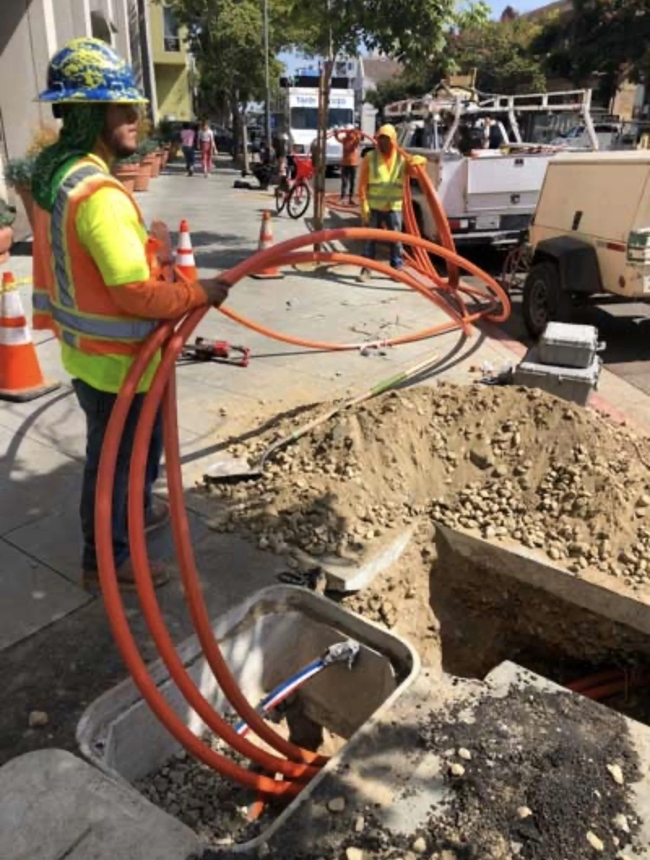 Technicians workers install underground  cables in a city sidewalk, with orange conduit pipes and a large excavation trench surrounded by construction equipment and traffic cones.