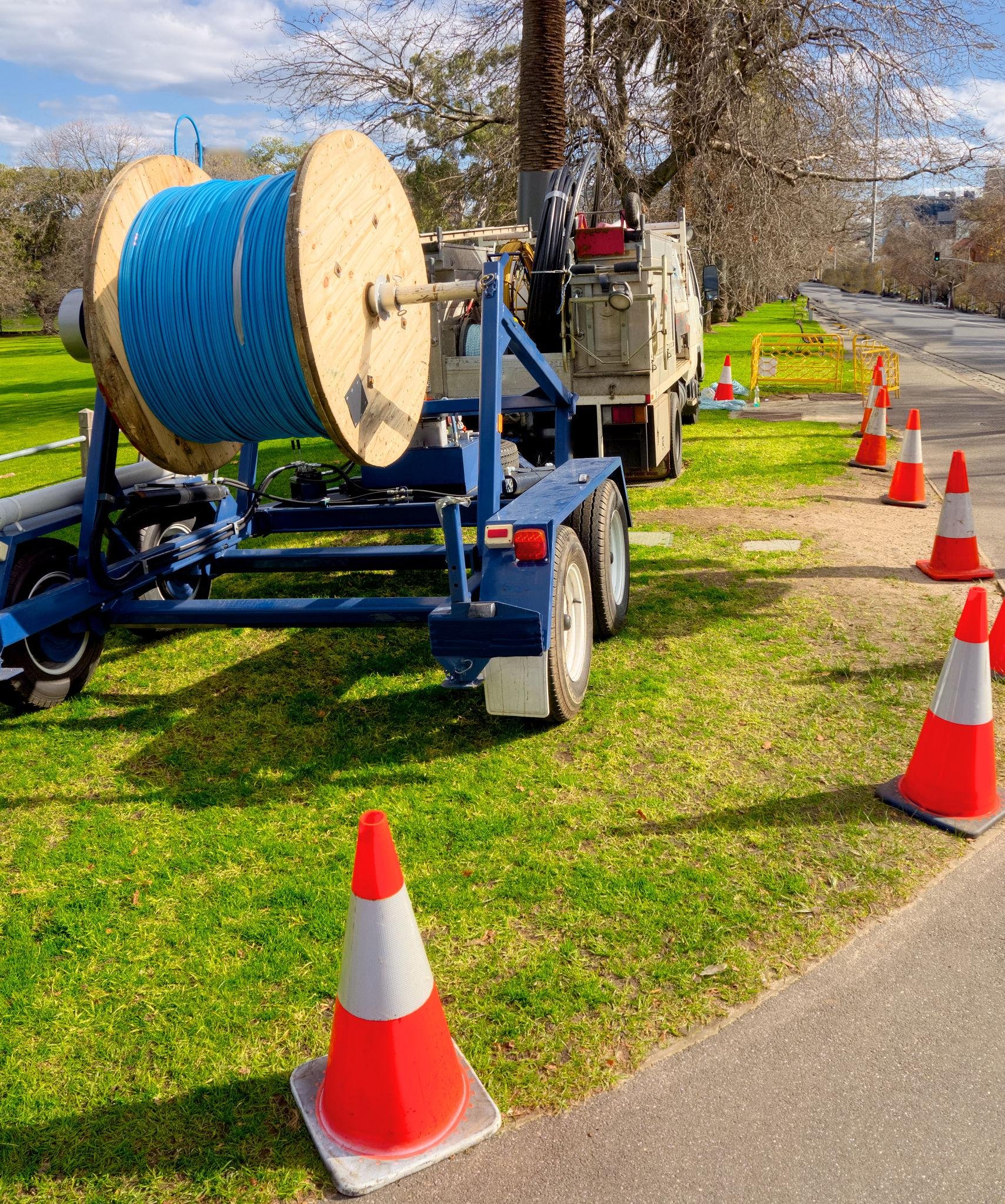 A street service vehicle is parked beside a grassy area during daytime, with orange traffic cones set up along the sidewalk. The vehicle has a large wooden cable spool with bright blue cable, and there are trees and a cityscape in the background.