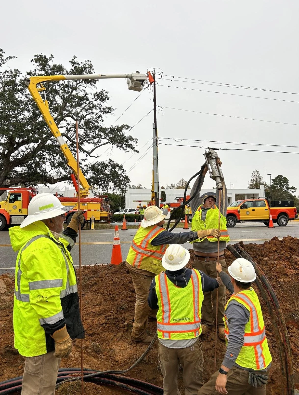 Workers in safety vests and hard hats repairing power lines with equipment and a cherry picker truck on a street.
