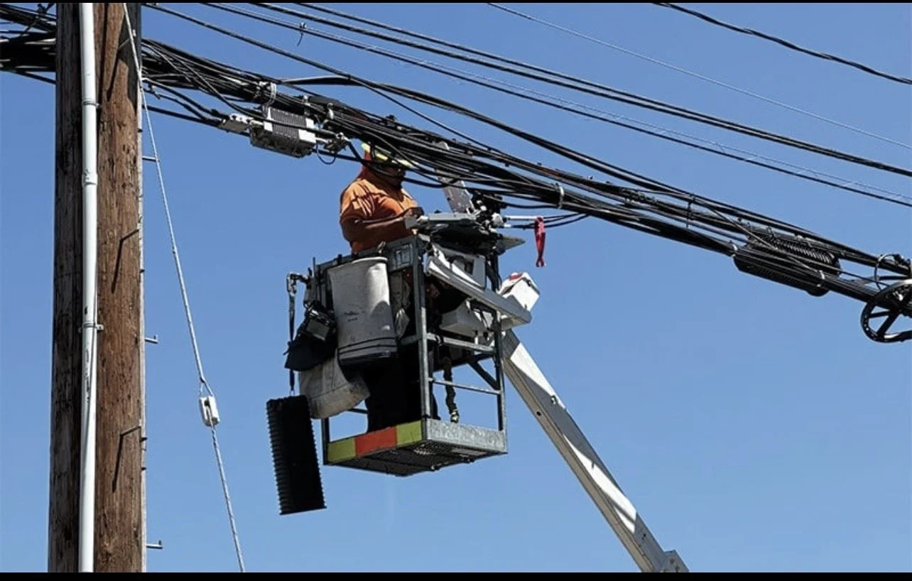 A utility worker on a bucket lift repairing electrical lines on a wooden utility pole against a clear blue sky.