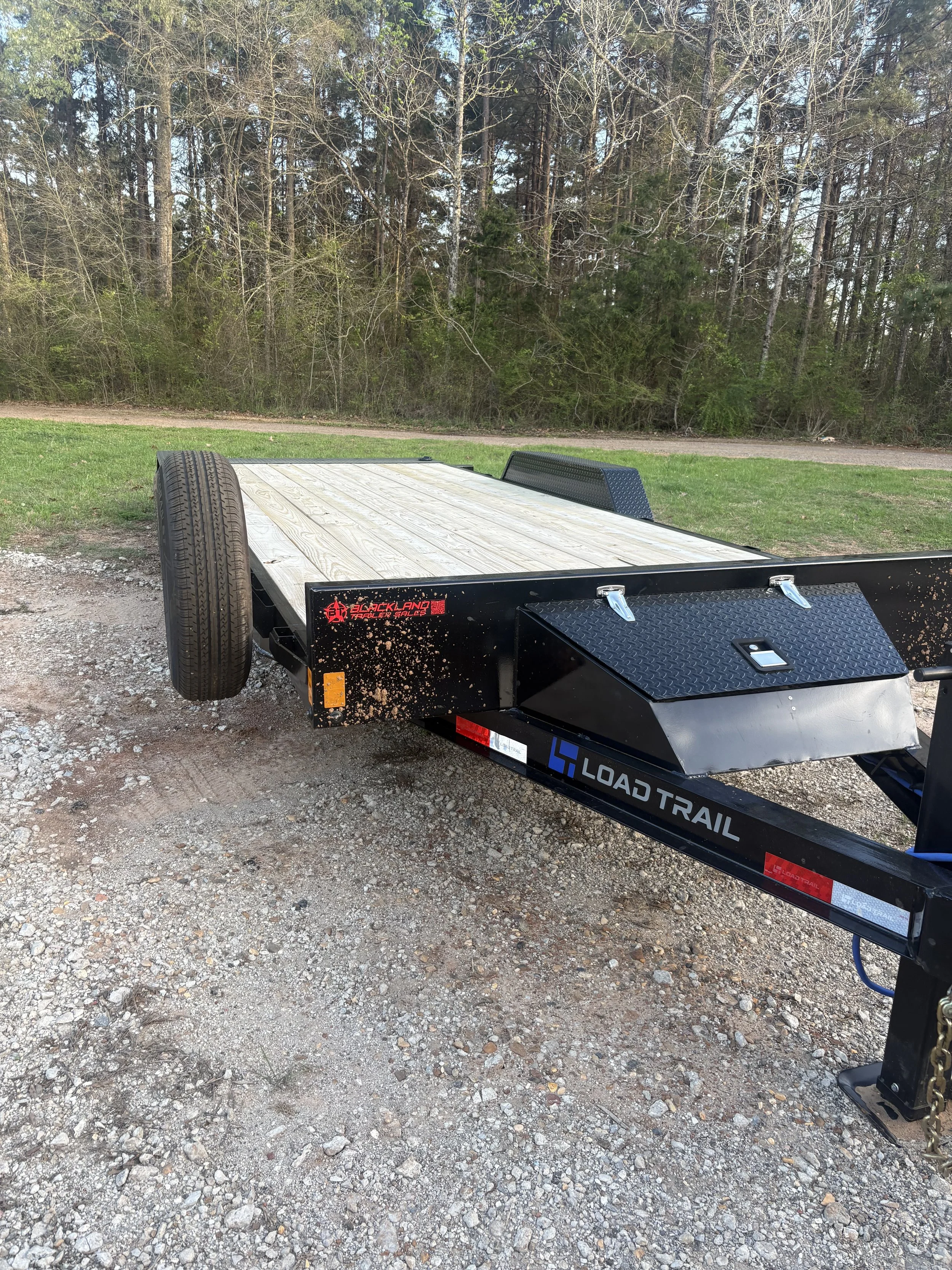 Black Load Trail flatbed trailer with wooden deck and a spare tire, parked on gravel with grass and trees in the background.
