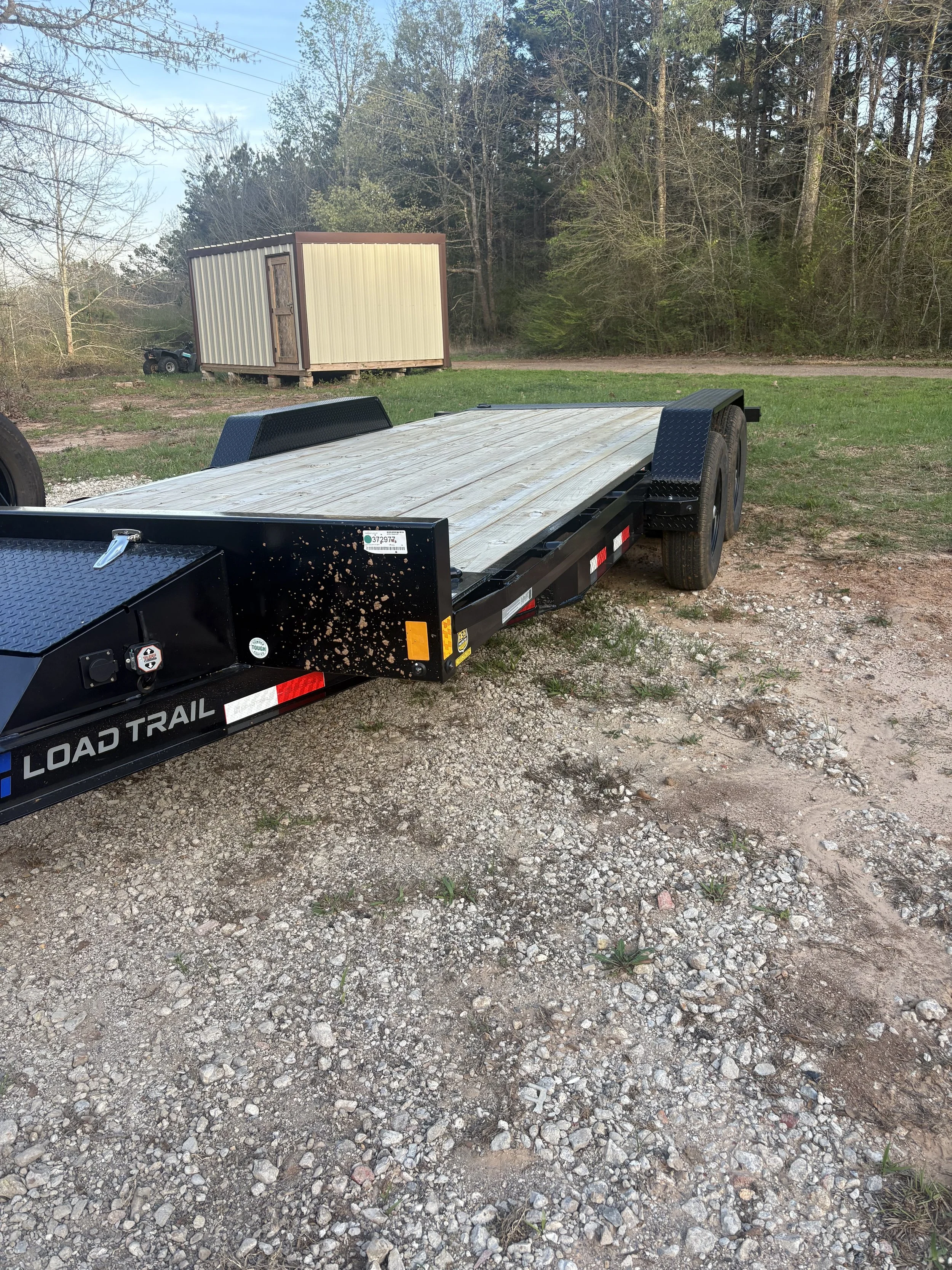 An empty black flatbed trailer with a wooden deck parked on a gravel surface with a small shed and forested area in the background.