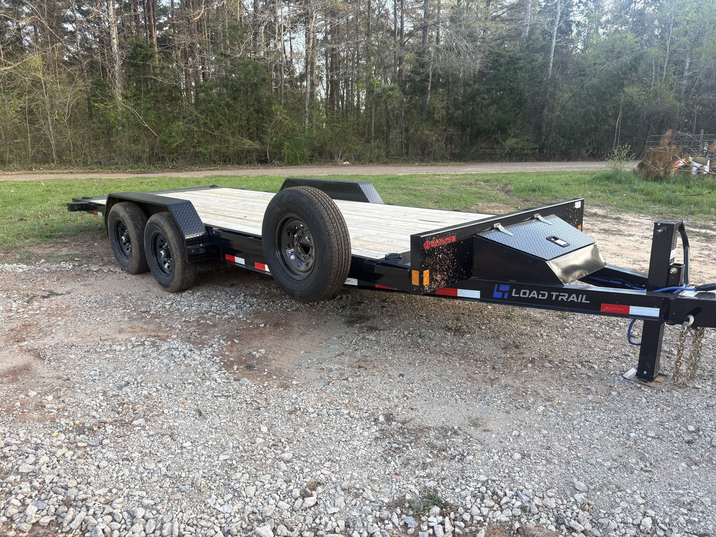 Black Load Trail flatbed trailer with dual axles and spare tire, parked on a dirt and gravel surface in front of a grassy area and wooded trees.