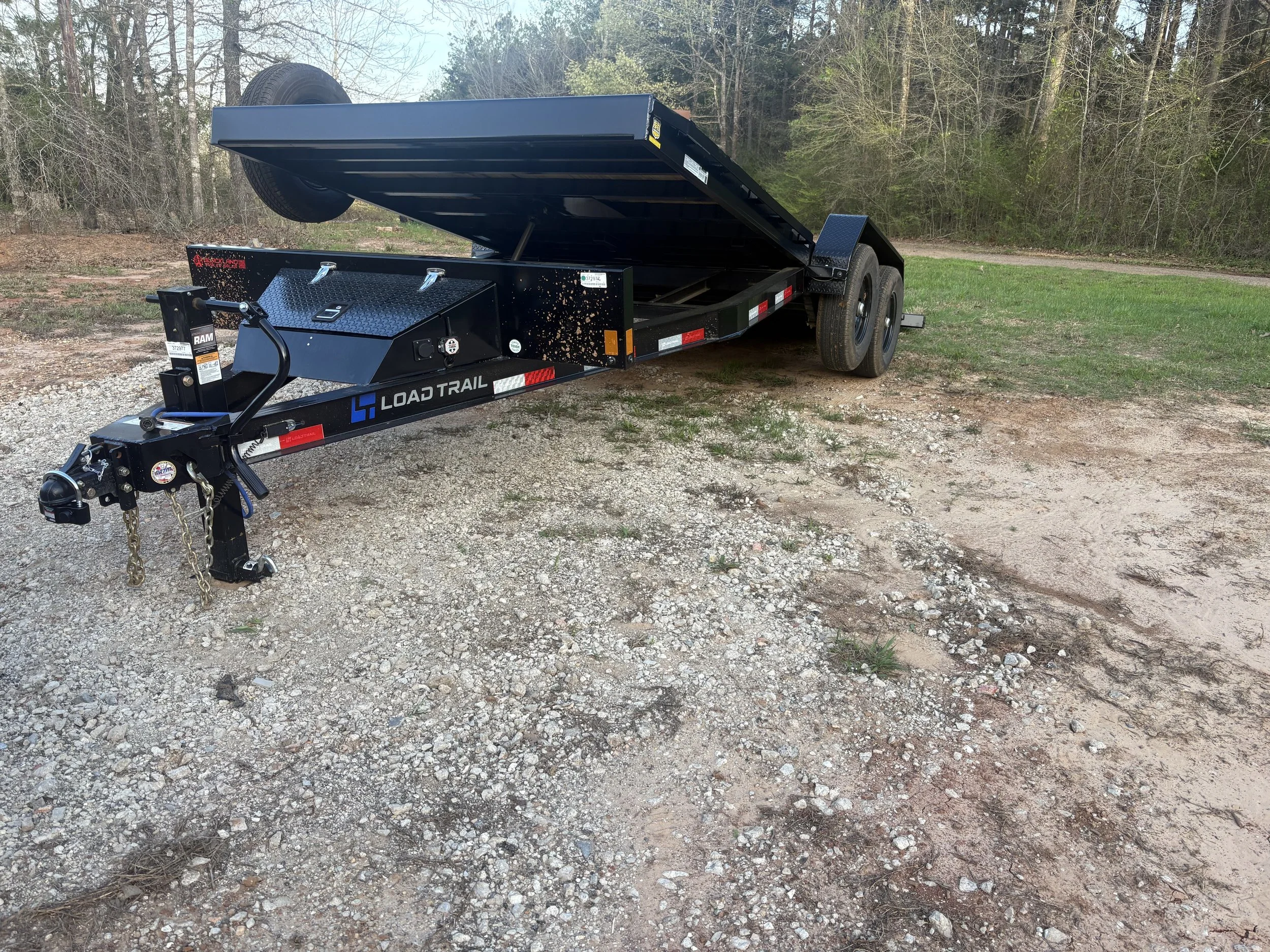 Black Load Trail flatbed trailer with a raised bed parked on a dirt patch with grass and trees in the background.