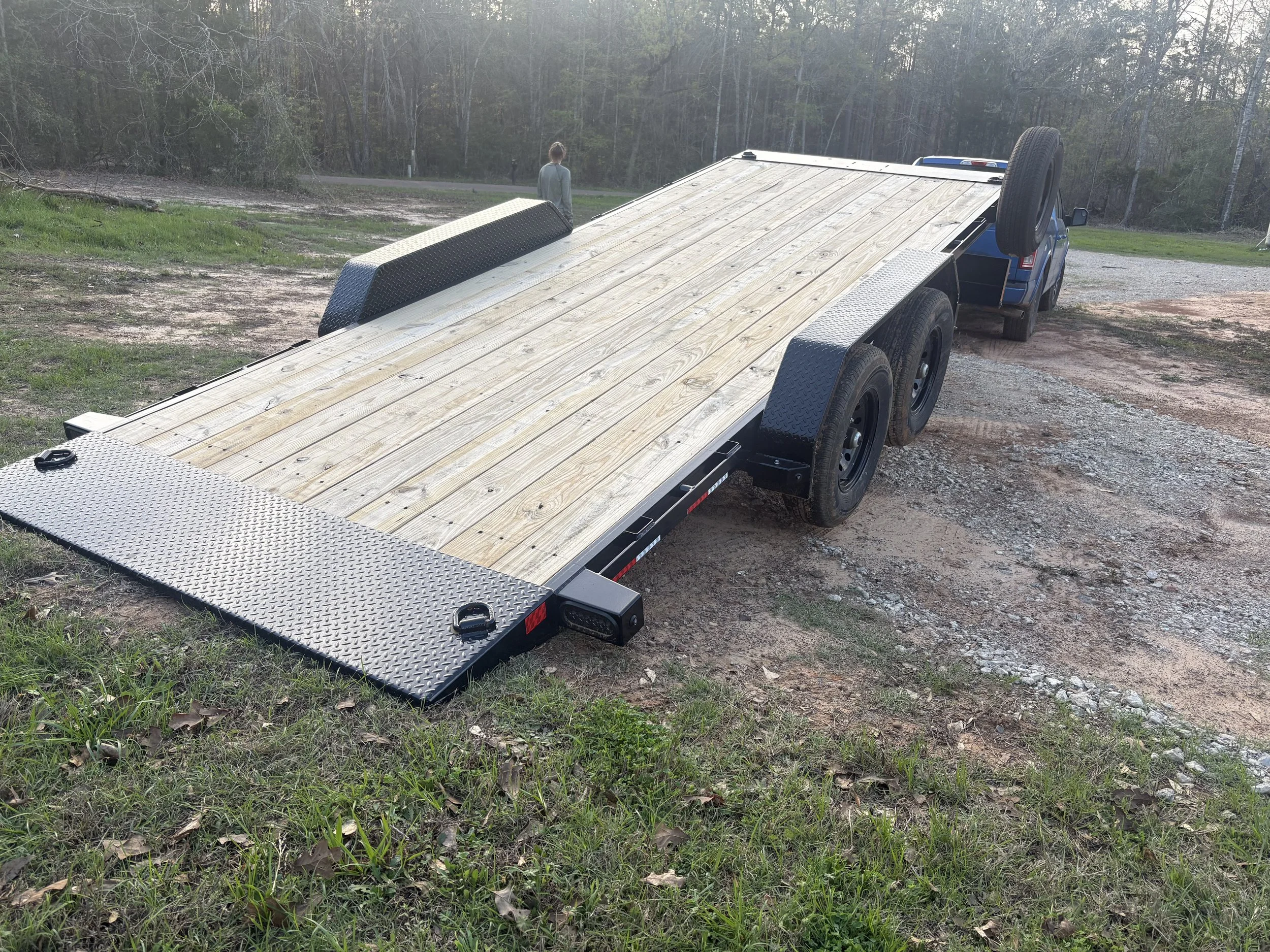 A flatbed trailer attached to a blue pickup truck, parked on a gravel and dirt area near some grass and trees.