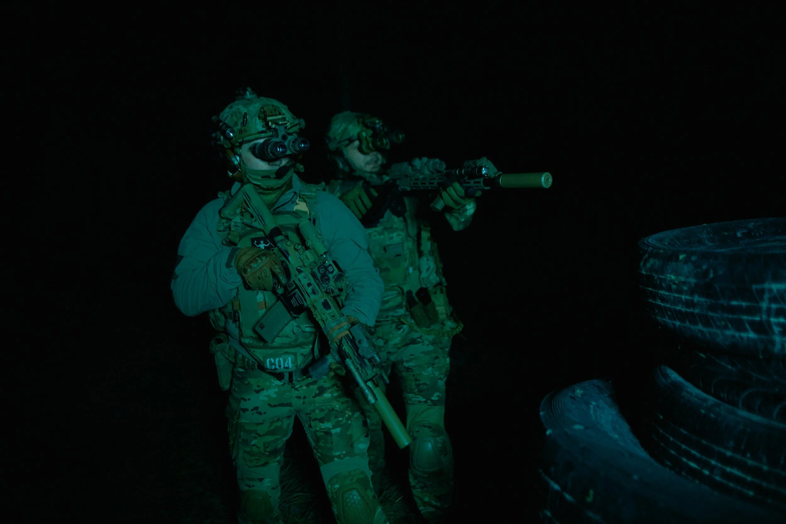 Two soldiers in camouflage gear with night vision goggles, holding rifles, in a dark environment with tires on the right side.