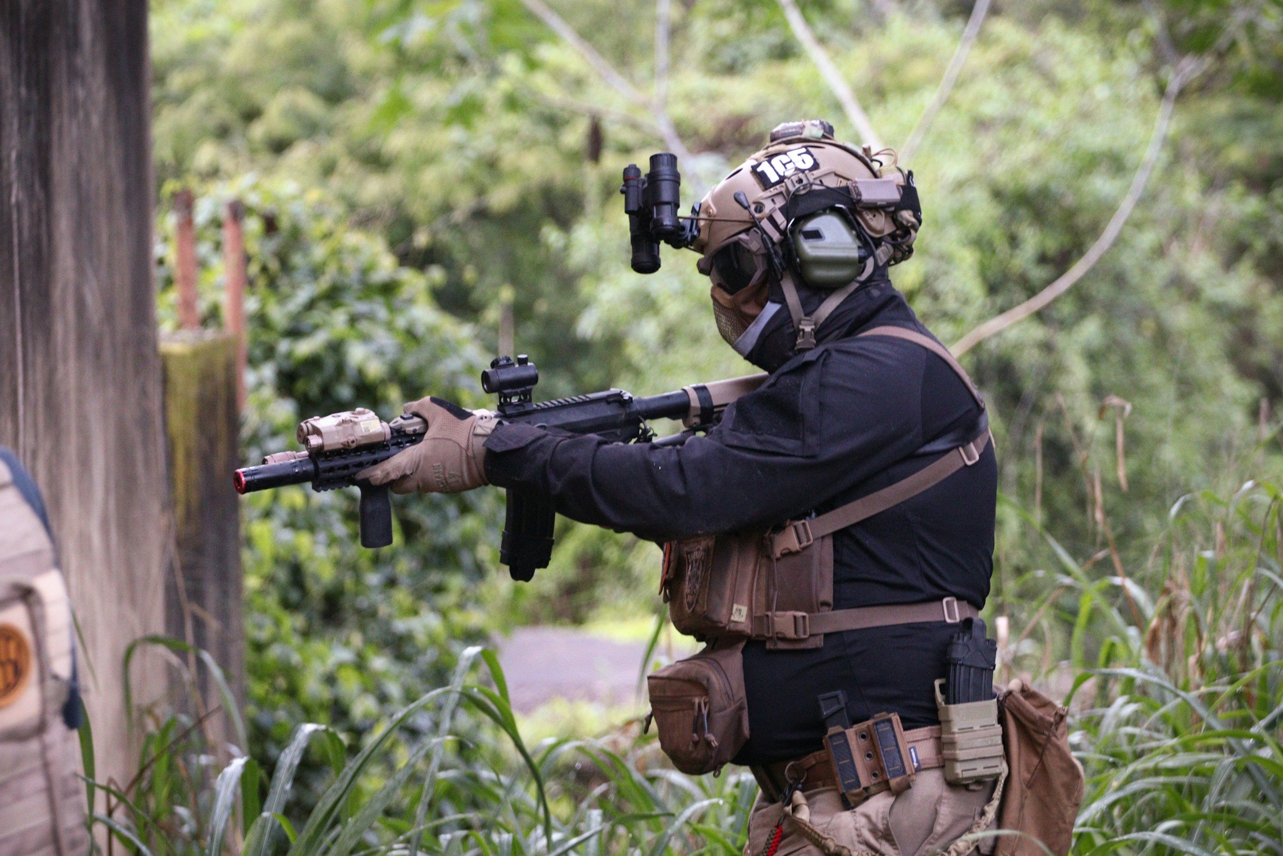 A person wearing tactical gear aiming a rifle in a forested area.