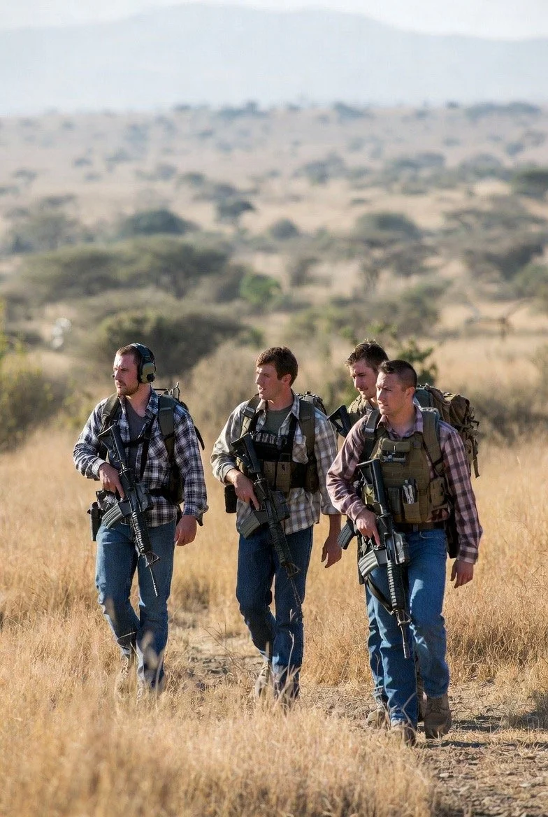 Four young men wearing plaid shirts, jeans, and backpacks walking across a dry field with rifles, in a rural landscape.