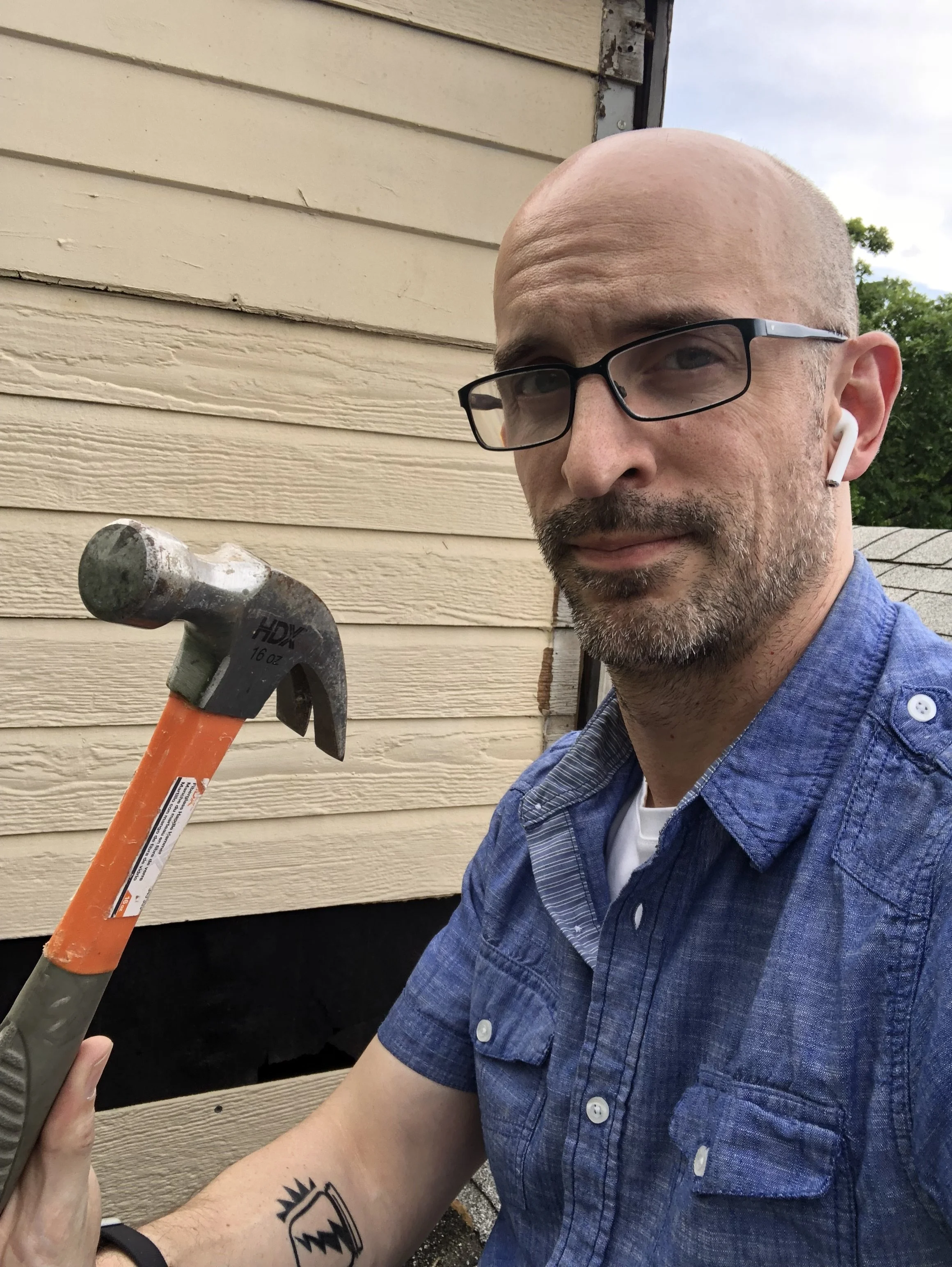 A man with glasses and AirPods holding a hammer, standing in front of a house with beige siding and a small section of damaged black and beige material on the wall.