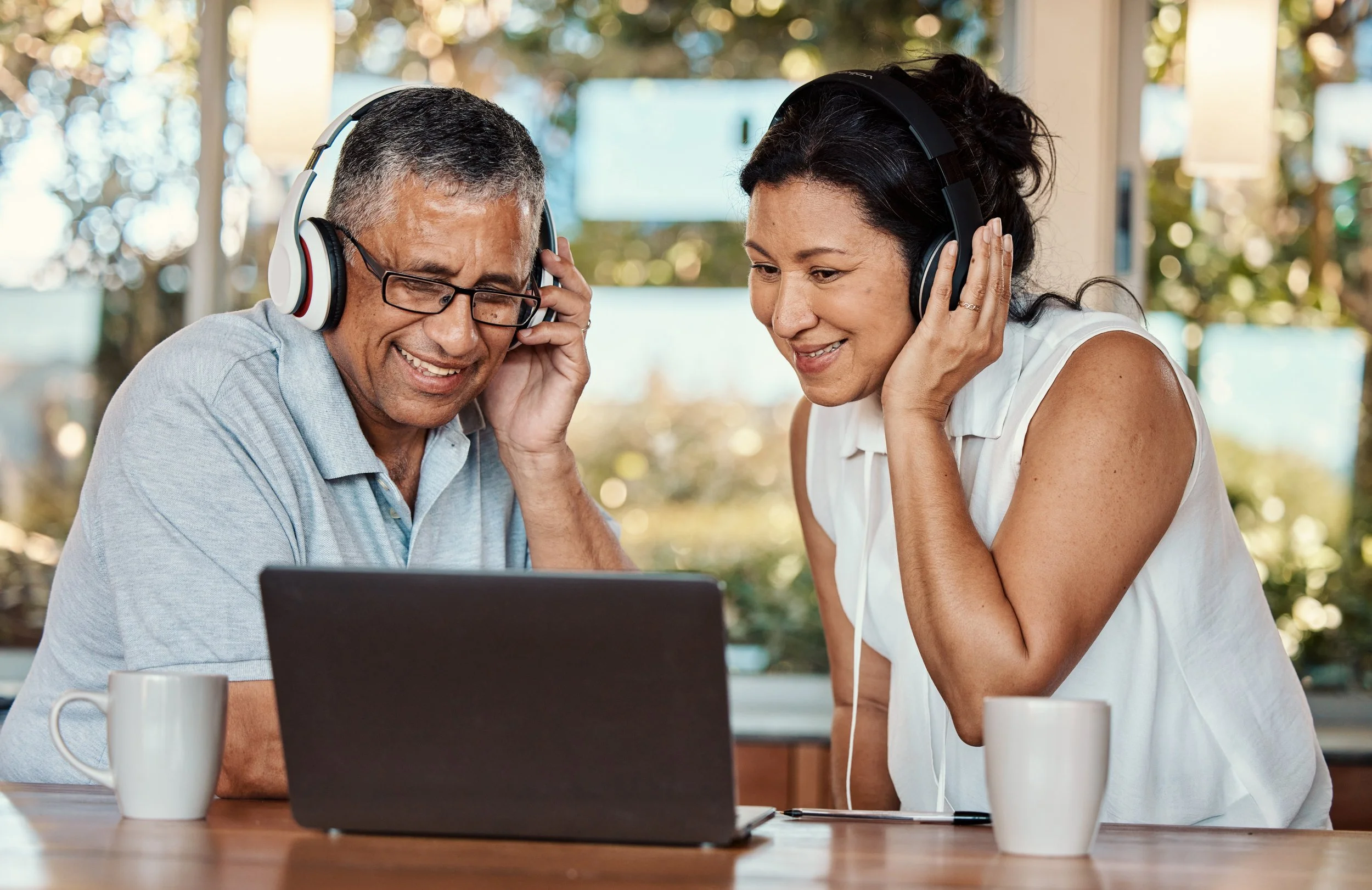 A man and woman wearing headphones looking at a laptop in a bright room with large windows and trees outside.