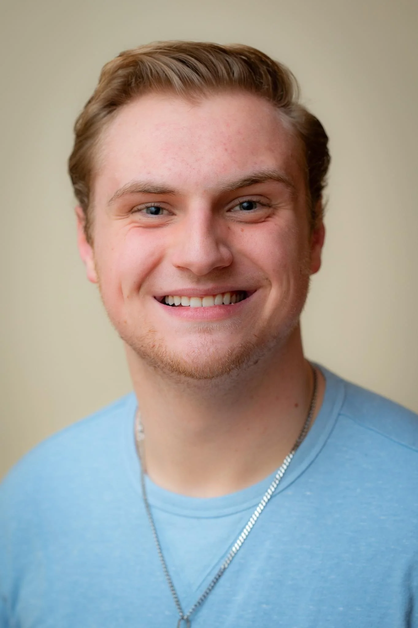 Close-up of a young man with light brown hair, blue eyes, smiling, wearing a light blue shirt, a silver chain necklace, and a zipper necklace, against a plain light-colored background.