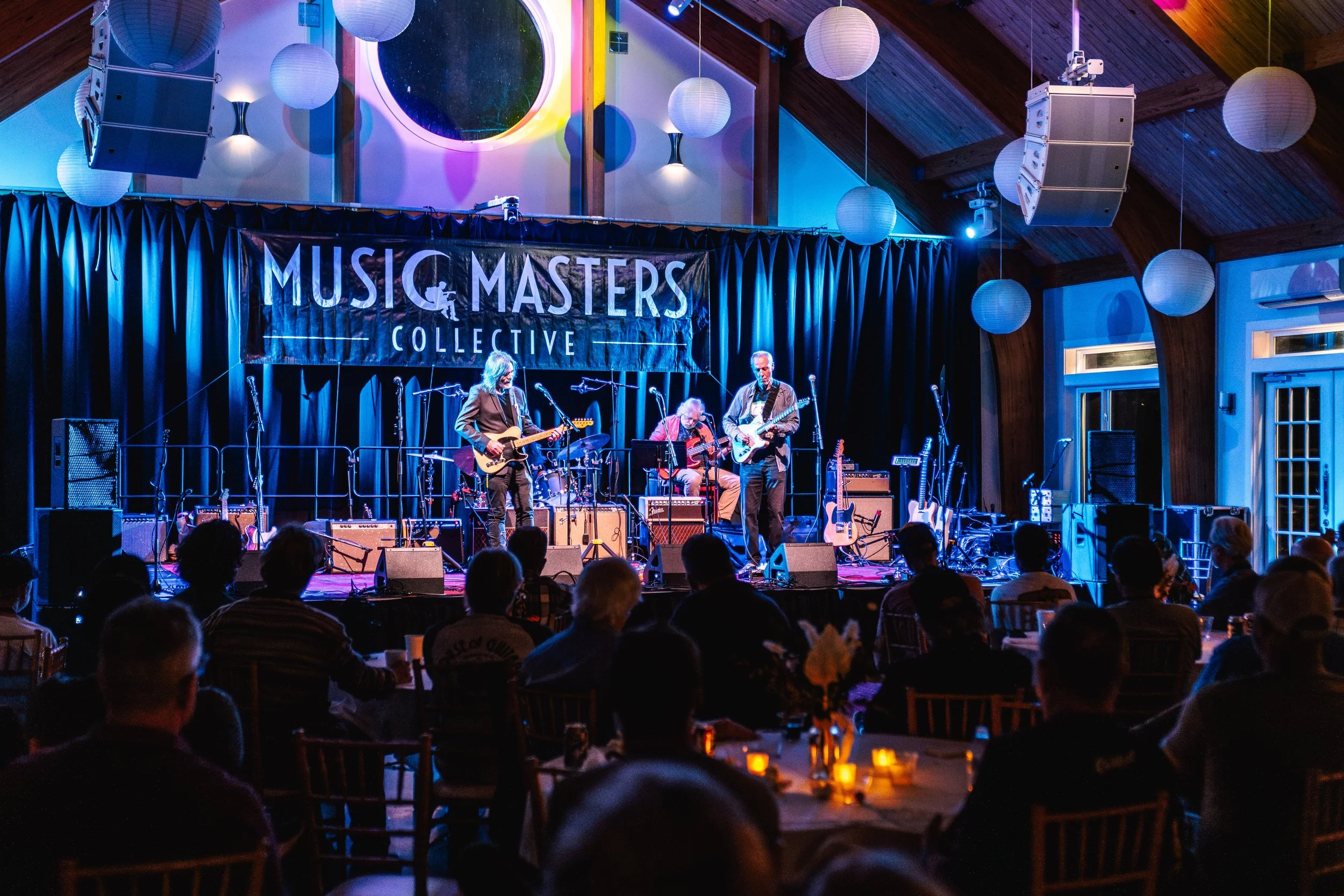 A band performing live on stage at a music event with a banner that reads 'Music Masters Collective'. The audience is seated at tables with lit candles, inside a wooden structure with round paper lanterns hanging from the ceiling.