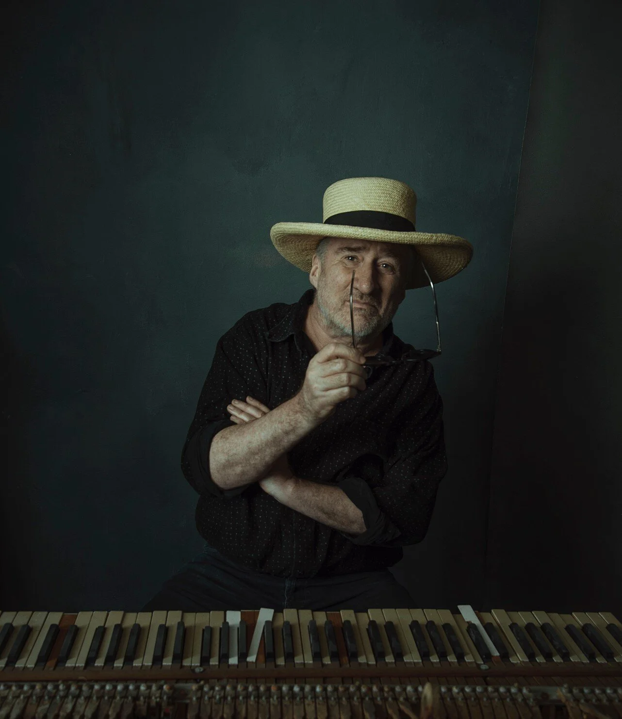 A man with a beard wearing a wide-brimmed straw hat and black shirt, sitting behind a piano with open lid, holding glasses in his right hand, with a dark green wall background.