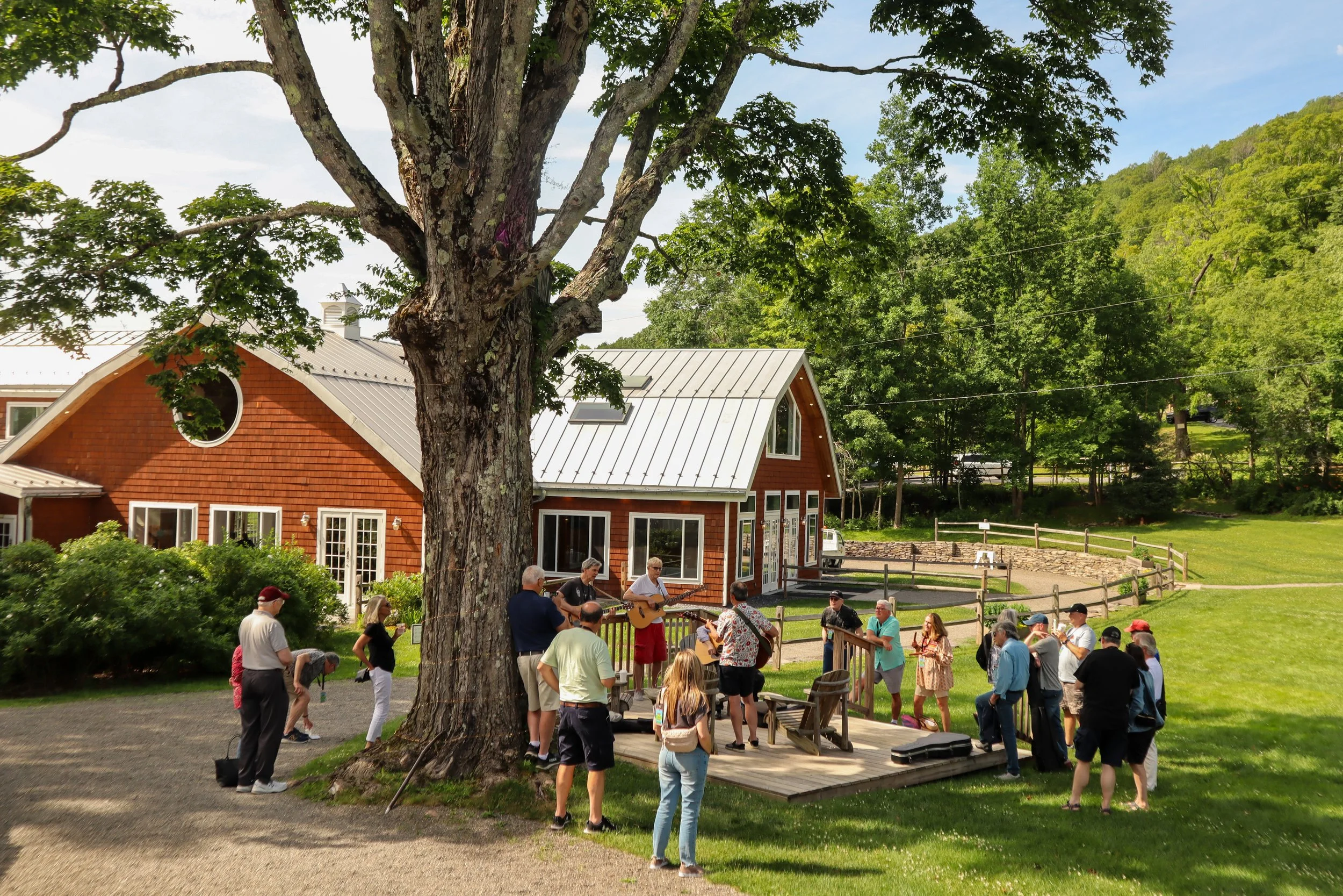 People gathered outdoors near a rustic building, enjoying live music on a small wooden stage with a guitar and microphone, under a large tree on a sunny day.