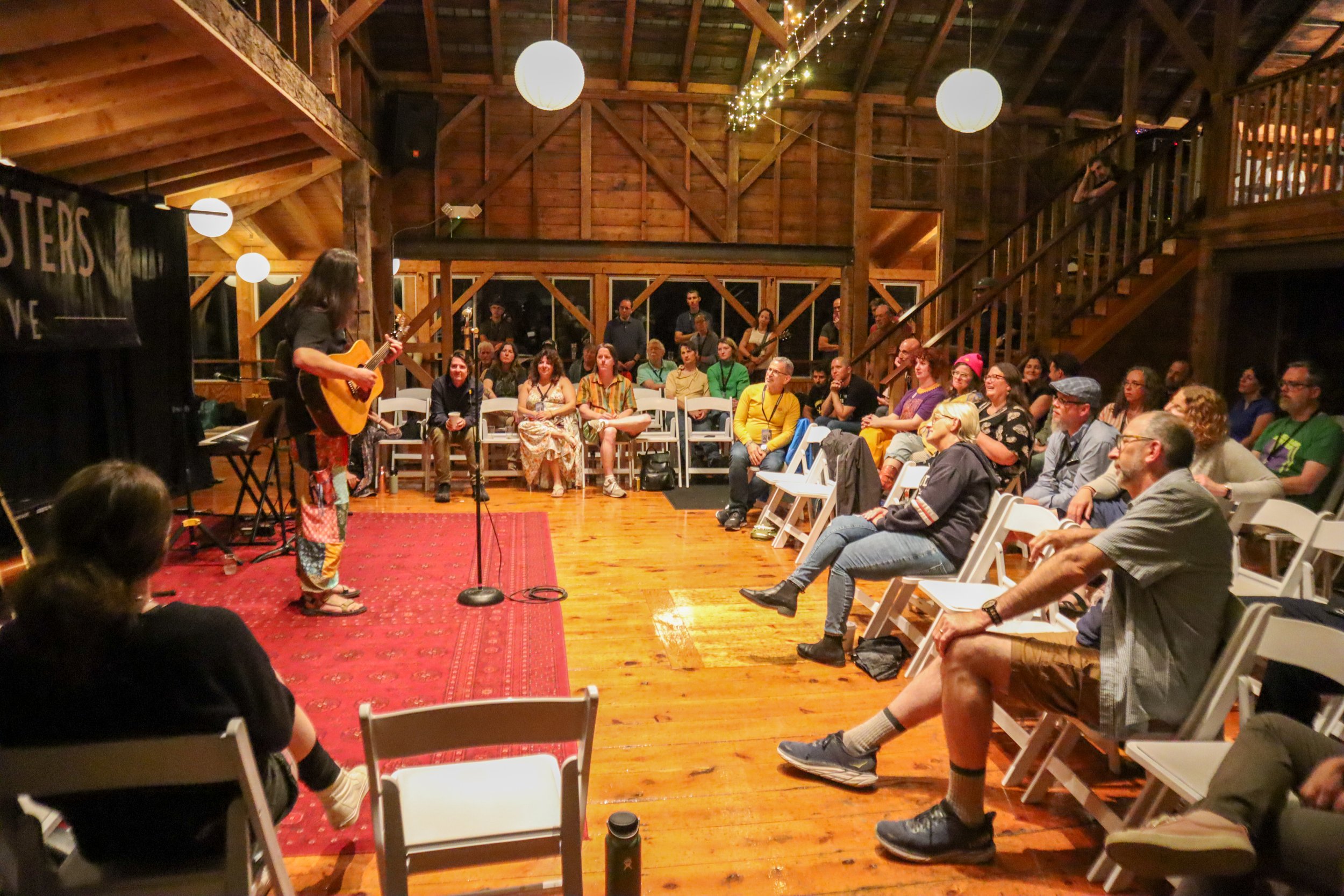 A person is performing with a guitar on a small stage in-front of an audience in a wooden venue during an intimate concert or event.