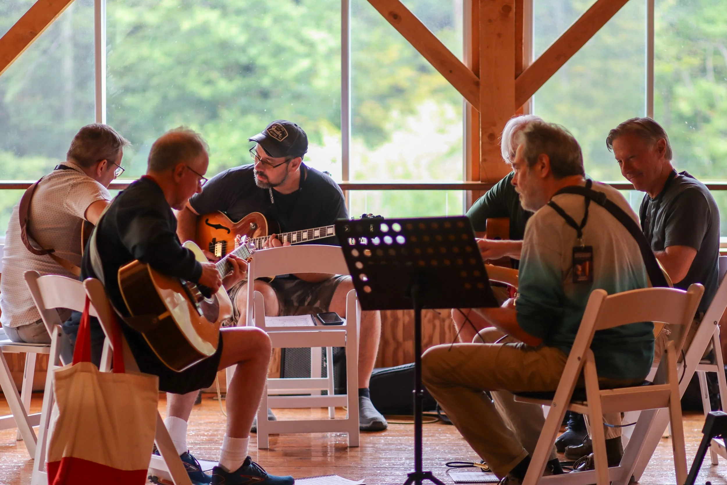 Group of people playing guitars and singing in a room with large windows and wooden beams.