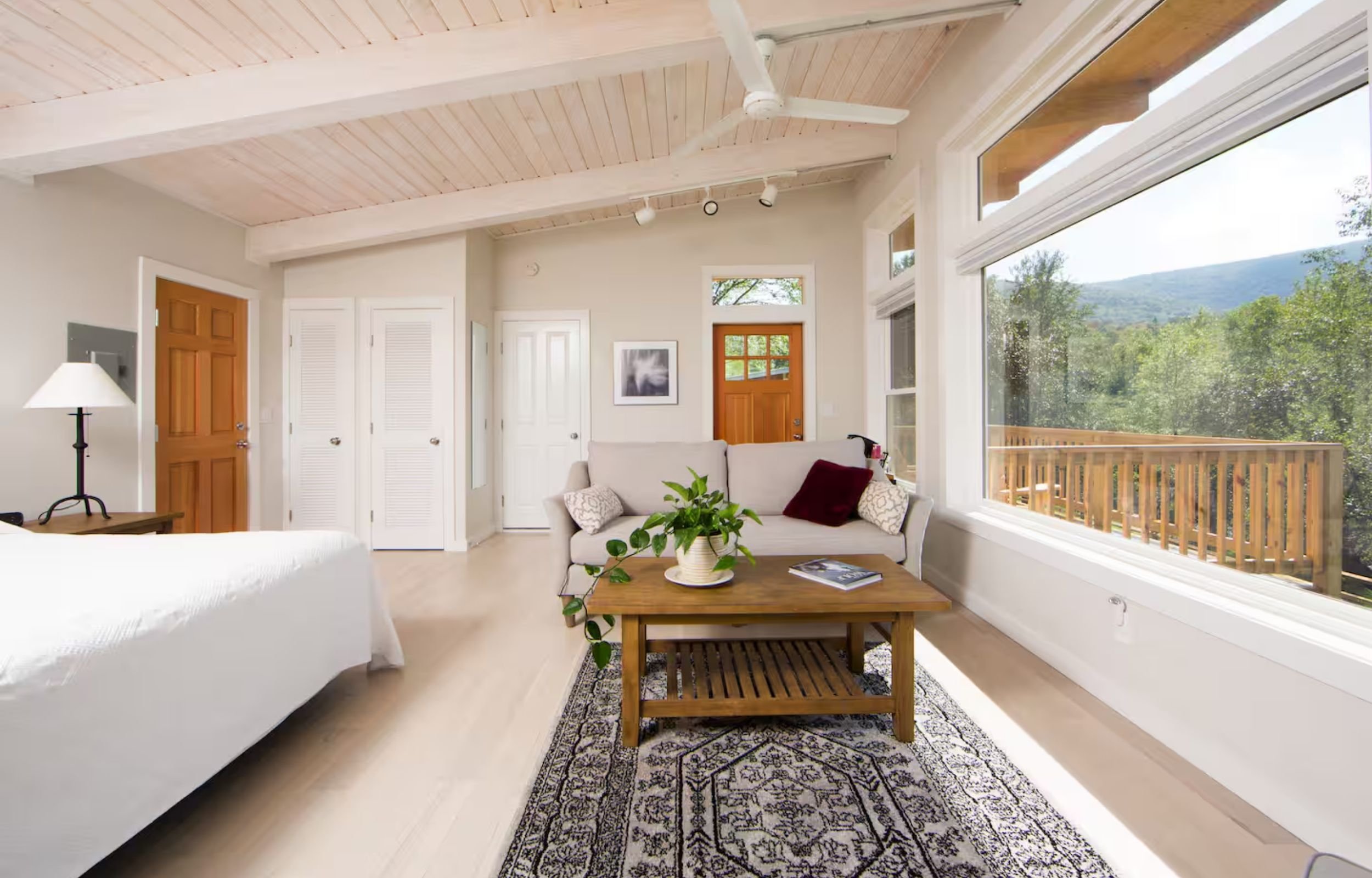 Bright living room with large windows revealing outdoor greenery, white walls, light wood flooring, white sofa with decorative pillows, wooden coffee table with a plant, area rug, and ceiling fan.
