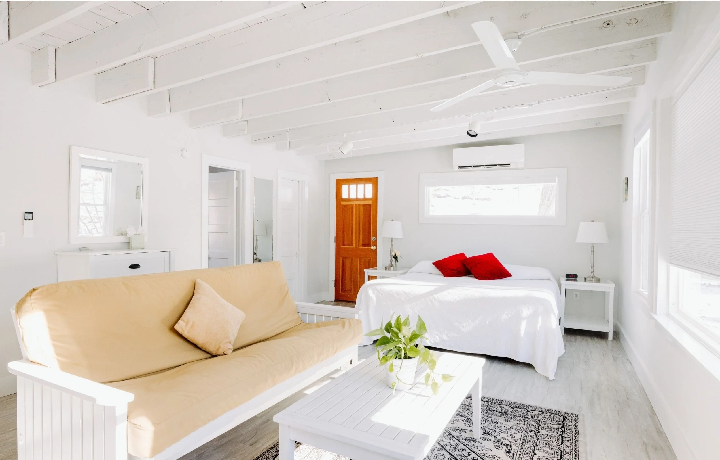 Bright, white living space with a bed in the background and a beige couch in the foreground, decorated with red and beige pillows, plants, and minimal furniture.