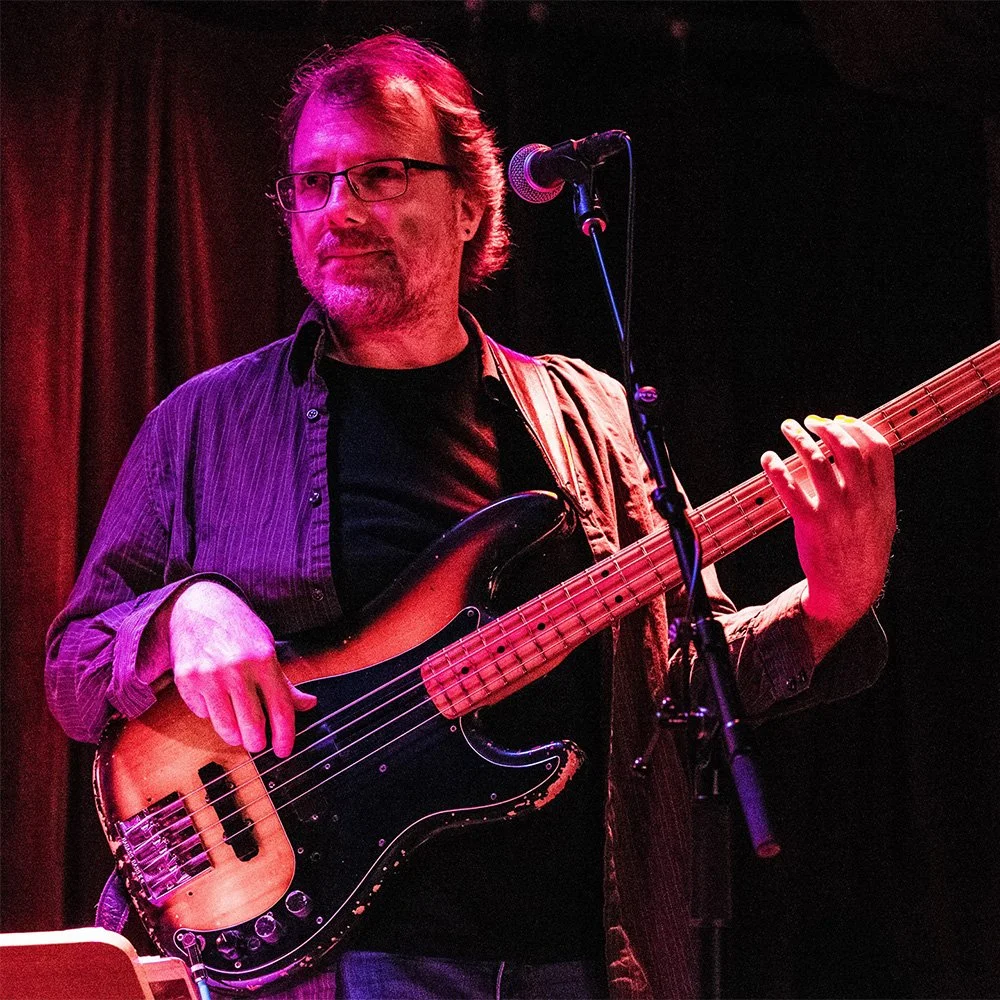 A man with glasses and a beard playing a bass guitar on stage with a microphone nearby, against a dark background with red curtains.