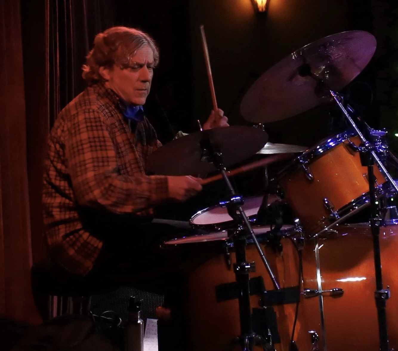 A man playing a drum set, wearing a plaid shirt, in a dimly lit venue.