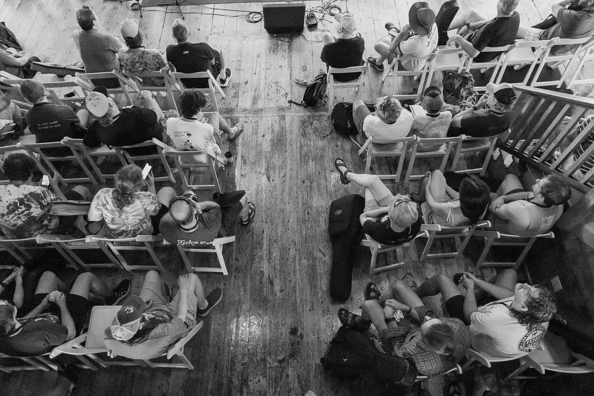 People sitting in a wooden-floored auditorium, facing towards a stage or screen, some using phones, dressed casually, with backpacks and hats visible.