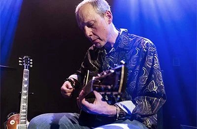 A man plays acoustic guitar on stage under blue lighting, with electric guitar set behind him.