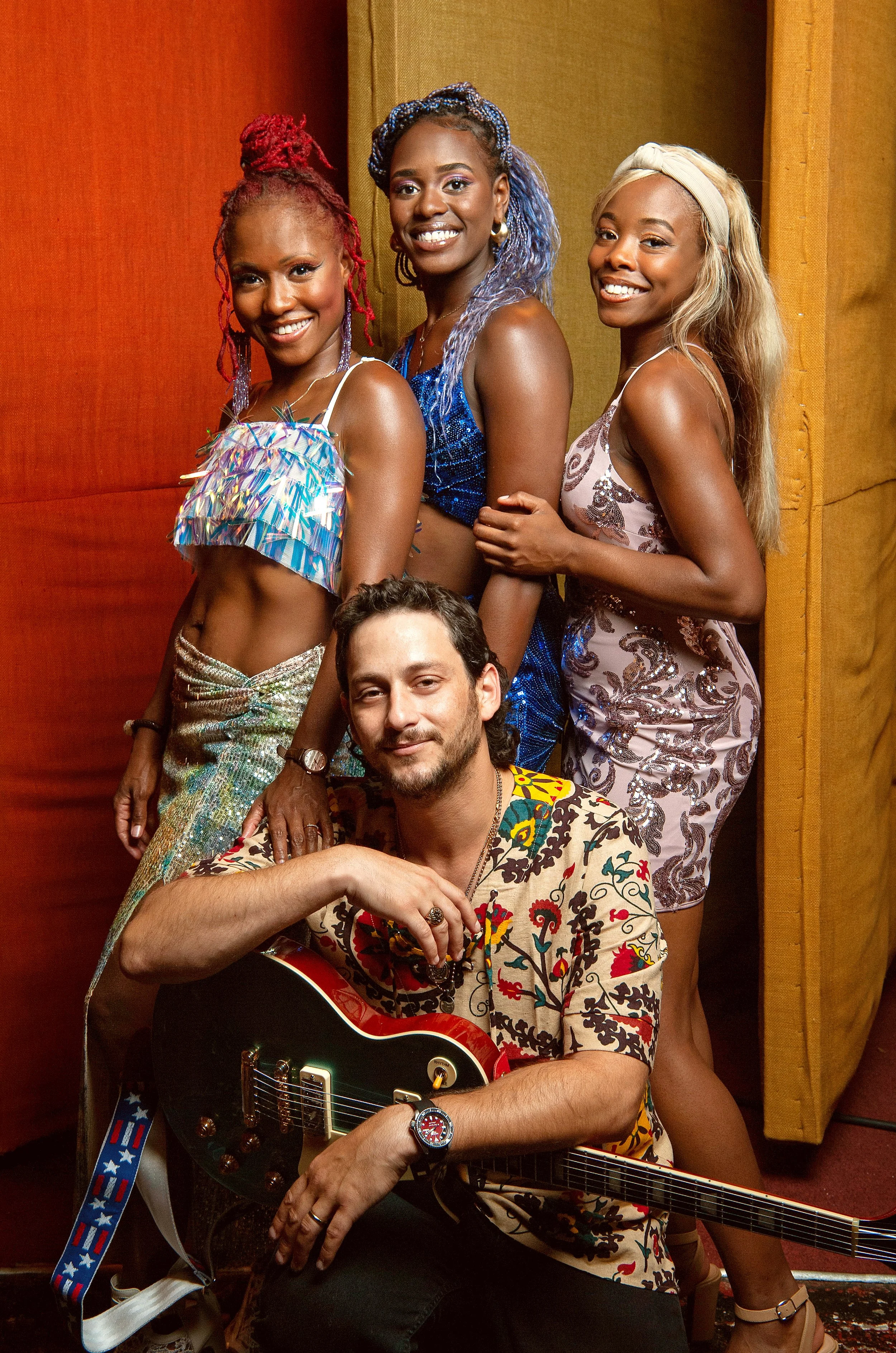 Five people, four women and one man, posing together indoors. The man is seated with a guitar, and the women stand behind him. They are dressed in colorful, patterned outfits and are smiling at the camera.