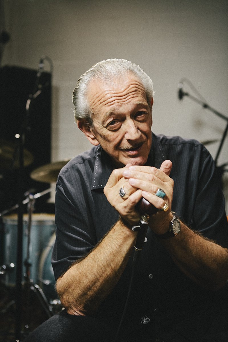 An elderly man with silver hair, wearing a black shirt, sitting in front of a microphone with drumming equipment in the background, smiling softly.