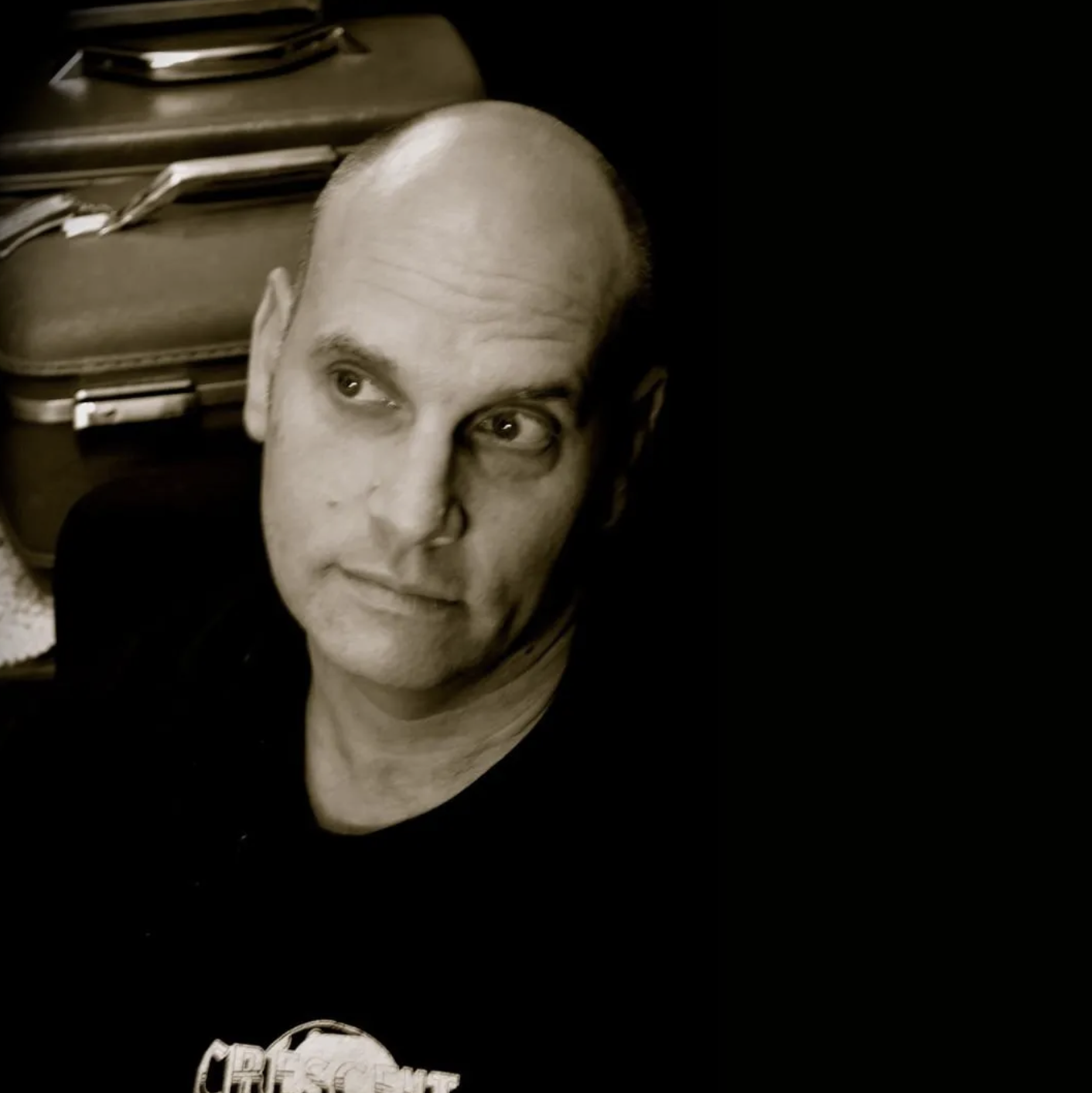 A man with a receding hairline looking to his left, with a serious expression, in front of a dark background and a brown briefcase.