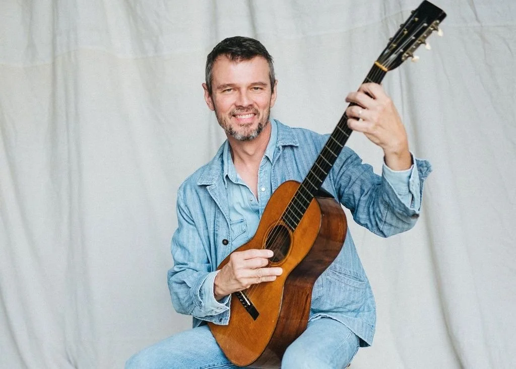 A man with a beard and short brown hair smiling while holding an acoustic guitar, wearing a denim shirt, sitting against a plain background.