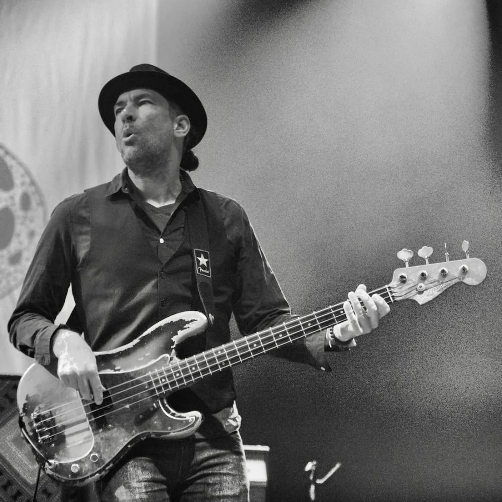 A man with a beard and mustache playing an electric bass guitar on stage. He is wearing a black hat, black shirt, and jeans, with a star patch on his shirt. The background shows a logo and stage equipment.