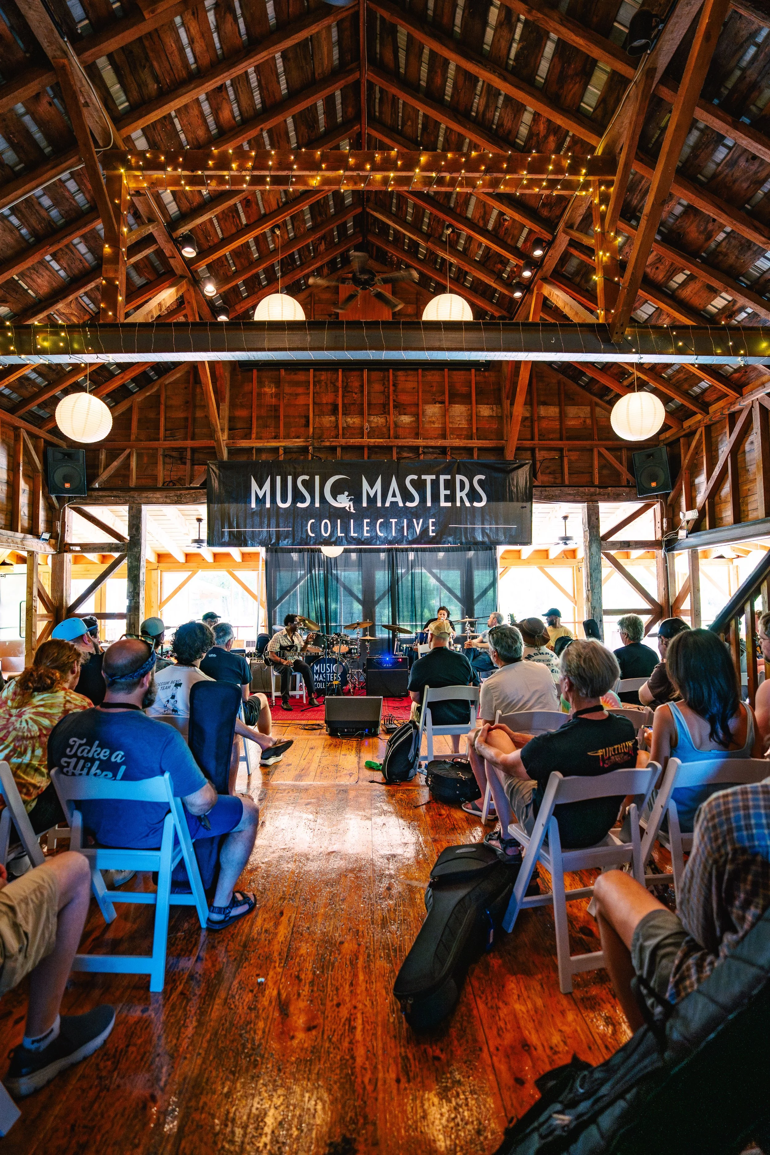 An indoor wooden concert venue with a high ceiling adorned with hanging paper lanterns and string lights, audience seated in chairs watching a band perform on stage under a large banner that reads 'Music Masters Collective.'