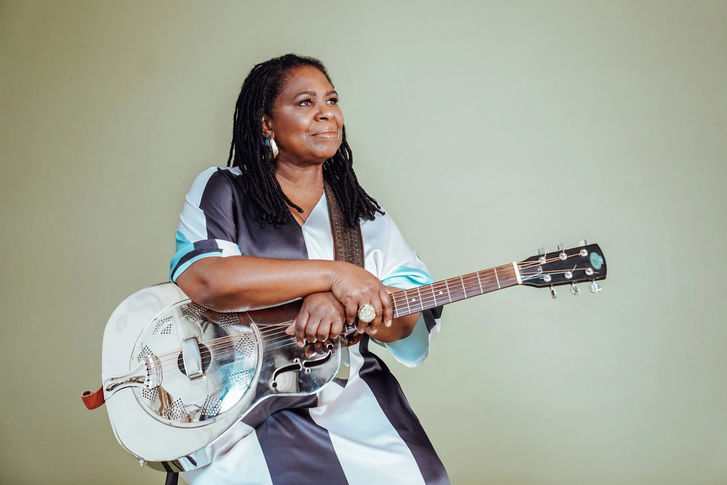 A woman with dark braided hair playing an acoustic guitar, sitting against a plain light green background.
