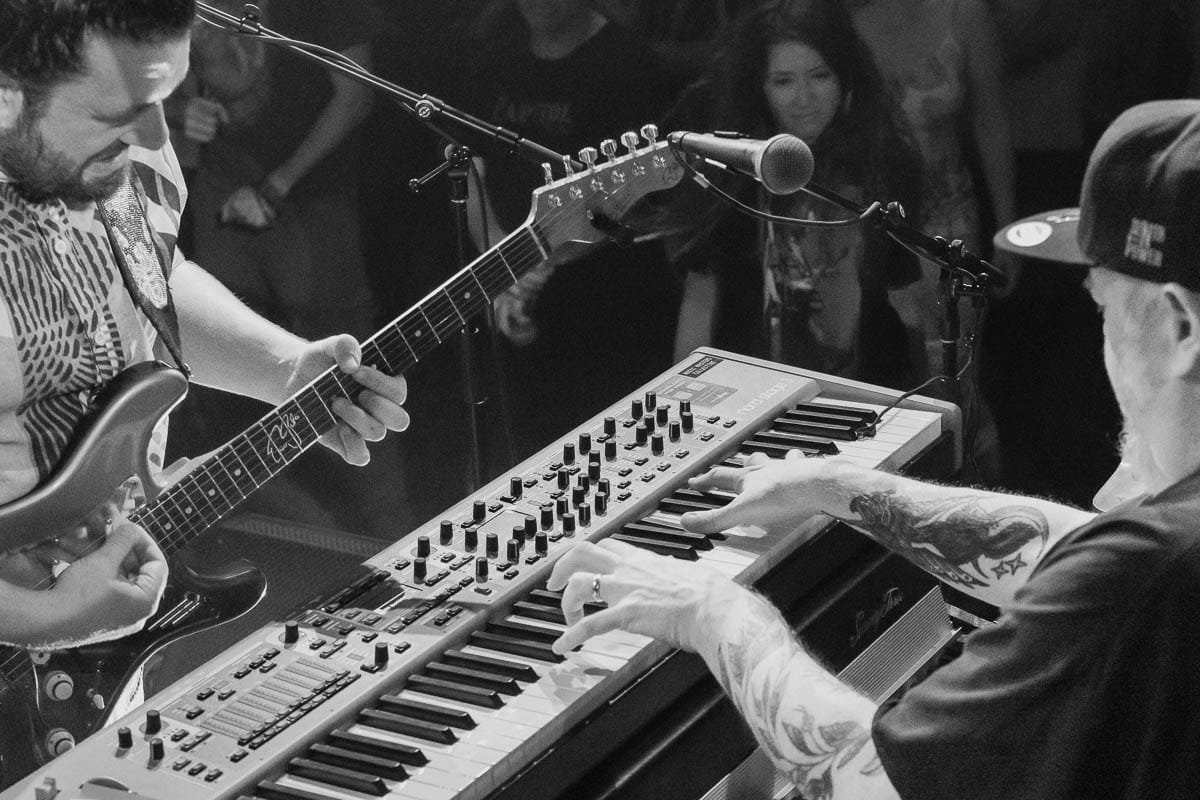 Musicians performing live music at a concert, with one playing an electric guitar and the other playing a keyboard, in front of an audience.