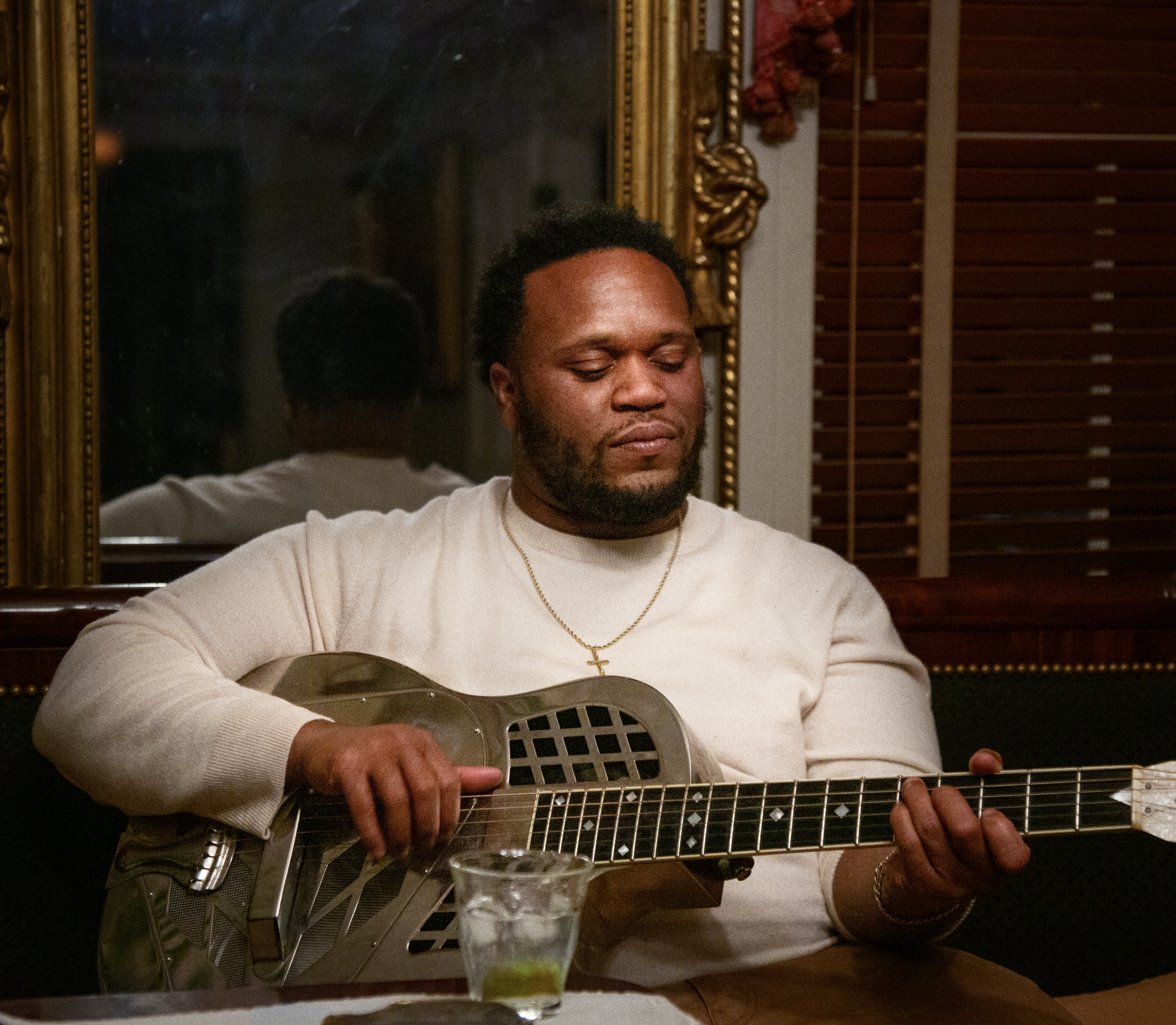 A man with a beard and short curly hair playing a resonator guitar in a warmly lit room, with a large ornate mirror behind him and wooden blinds on the window.