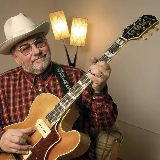 A man wearing glasses, a white fedora, and a red plaid shirt playing an acoustic guitar in a cozy room with a floor lamp in the background.