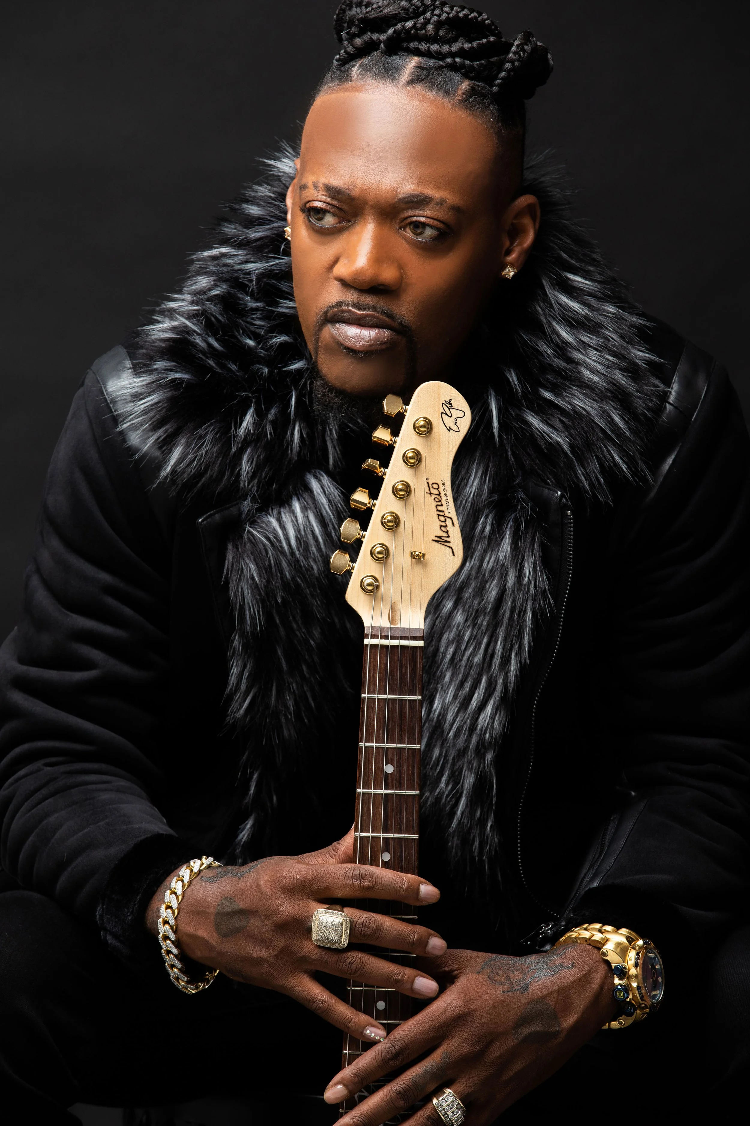A man with braided hair and facial hair, wearing a black jacket with faux fur collar, holds a guitar with a natural wood finish, signed by the artist, against a black background.