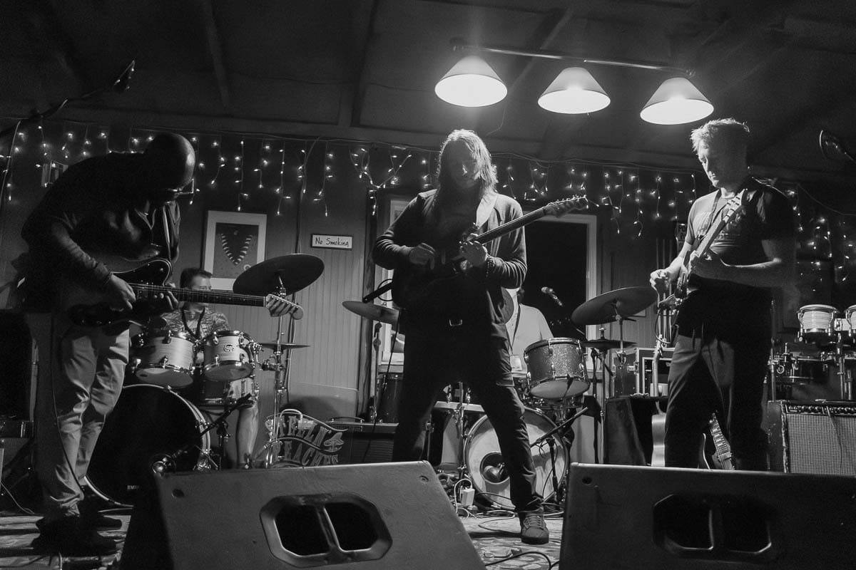 A band of five musicians performs on a stage with string lights in the background. The band includes a drummer, two guitarists, a keyboardist, and one more musician partly visible. The scene is in black and white.