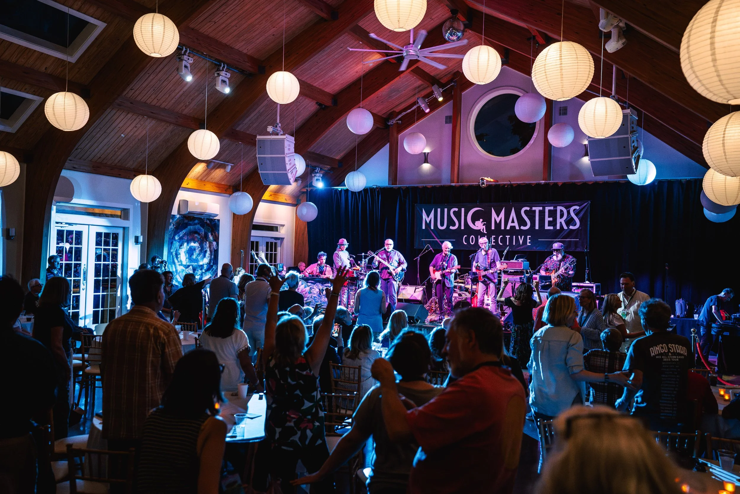 Concert at an indoor venue with a band performing on stage, called 'Music Masters Collective,' while the audience dances and enjoys the live music under hanging paper lanterns in a wooden-roofed hall.