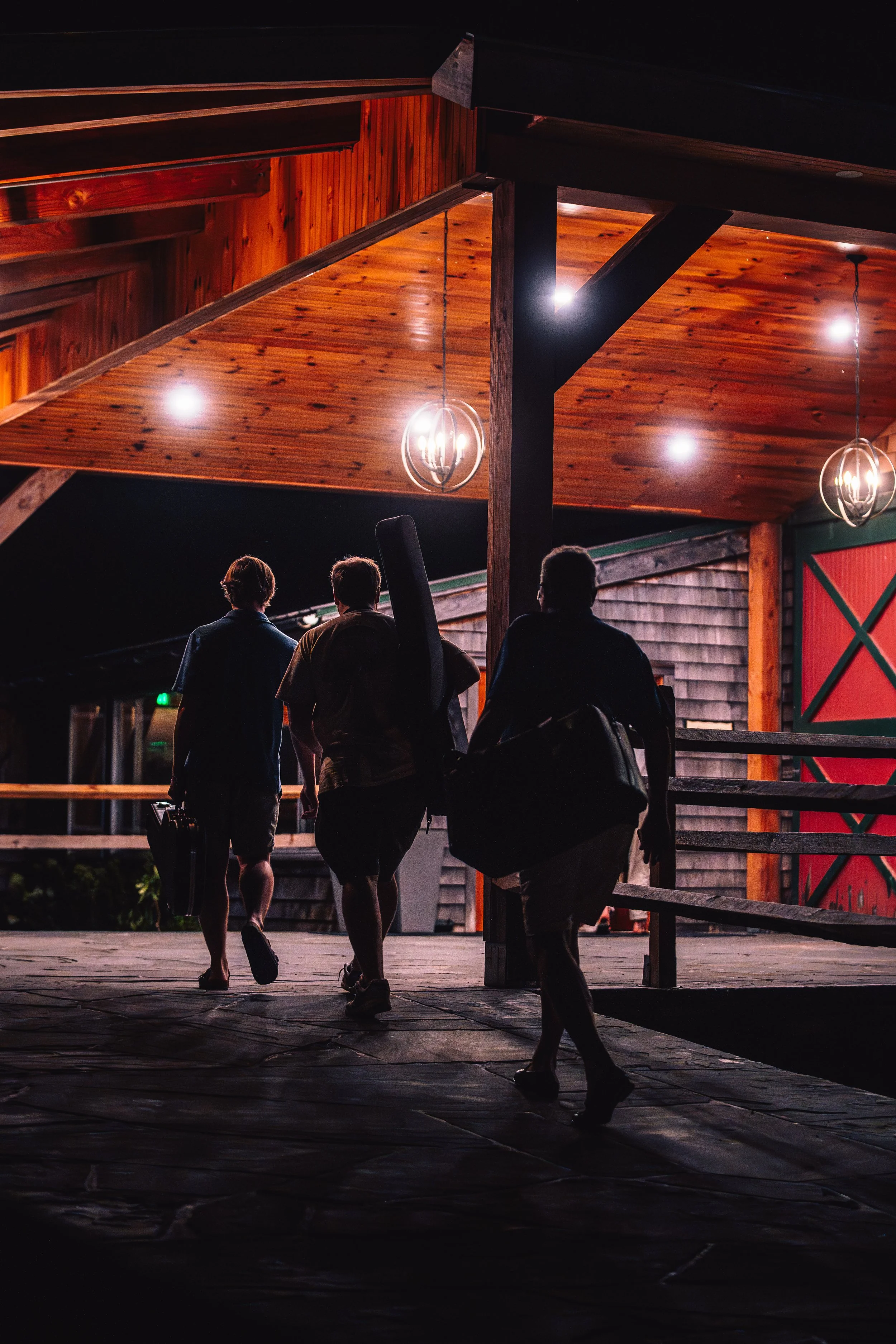 Three people walking into a wooden building at night, carrying guitar cases. The building has a sloped wooden ceiling with hanging lights.