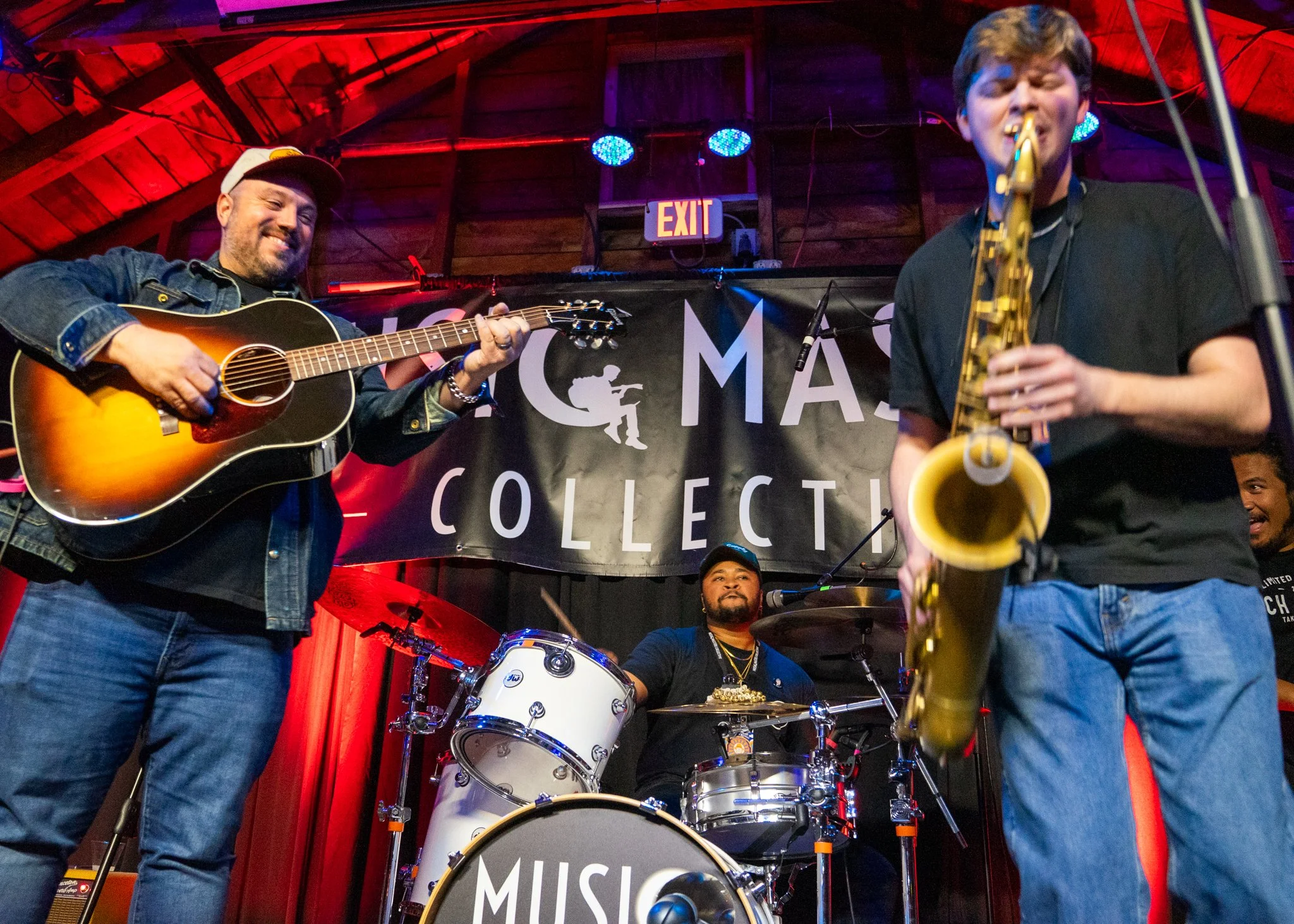 Musicians performing on stage with a banner in the background that reads 'Music Collection.' The band includes a guitarist, a drummer, and a saxophonist.