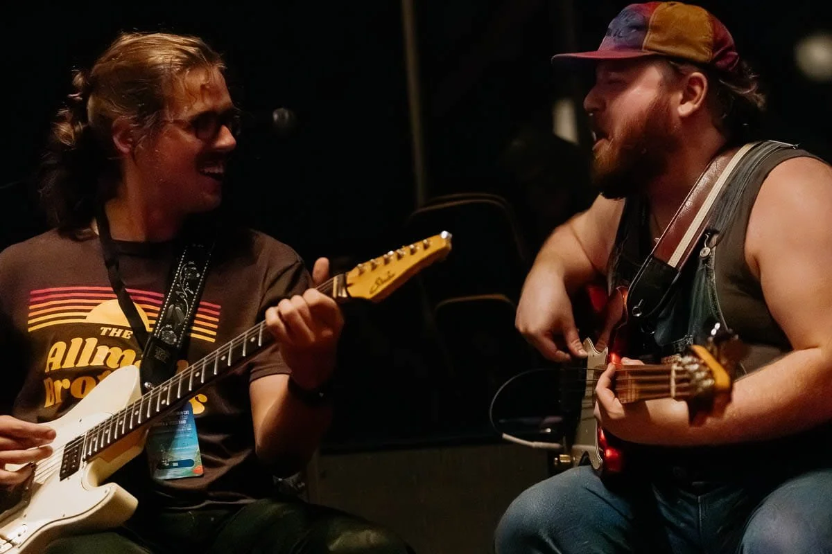 Two musicians, a woman and a man, are playing guitars and smiling at each other during a performance.