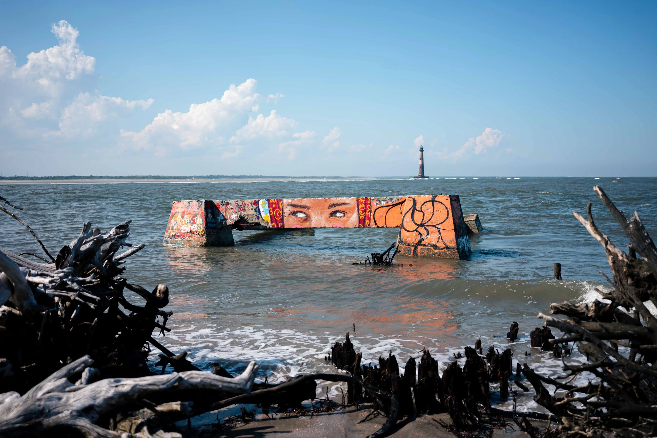 Graffiti-covered concrete structure in the ocean with a lighthouse in the background under a partly cloudy sky.