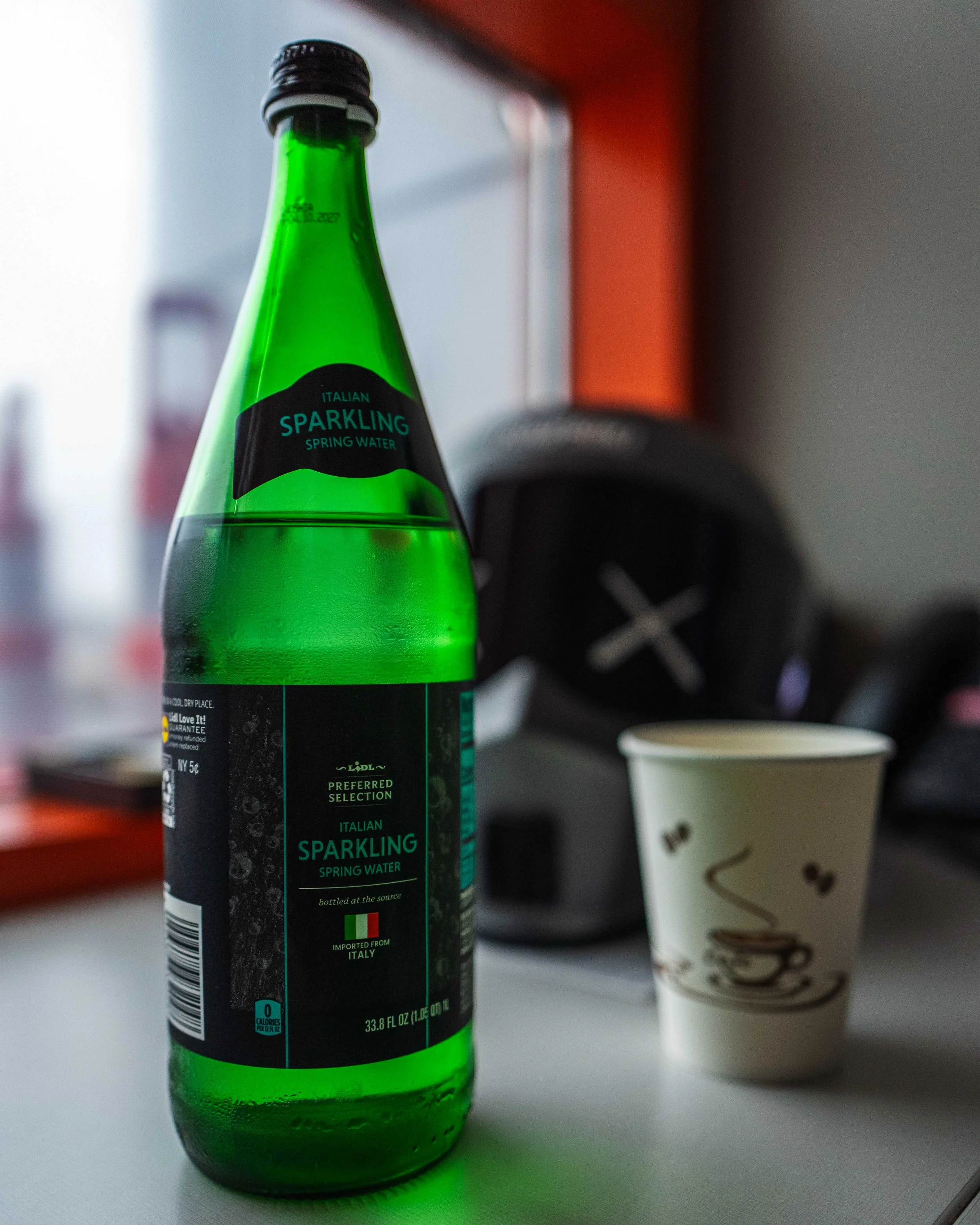 Green bottle of Italian sparkling spring water on a white surface, with a coffee cup and blurred electronic device in the background near a window.